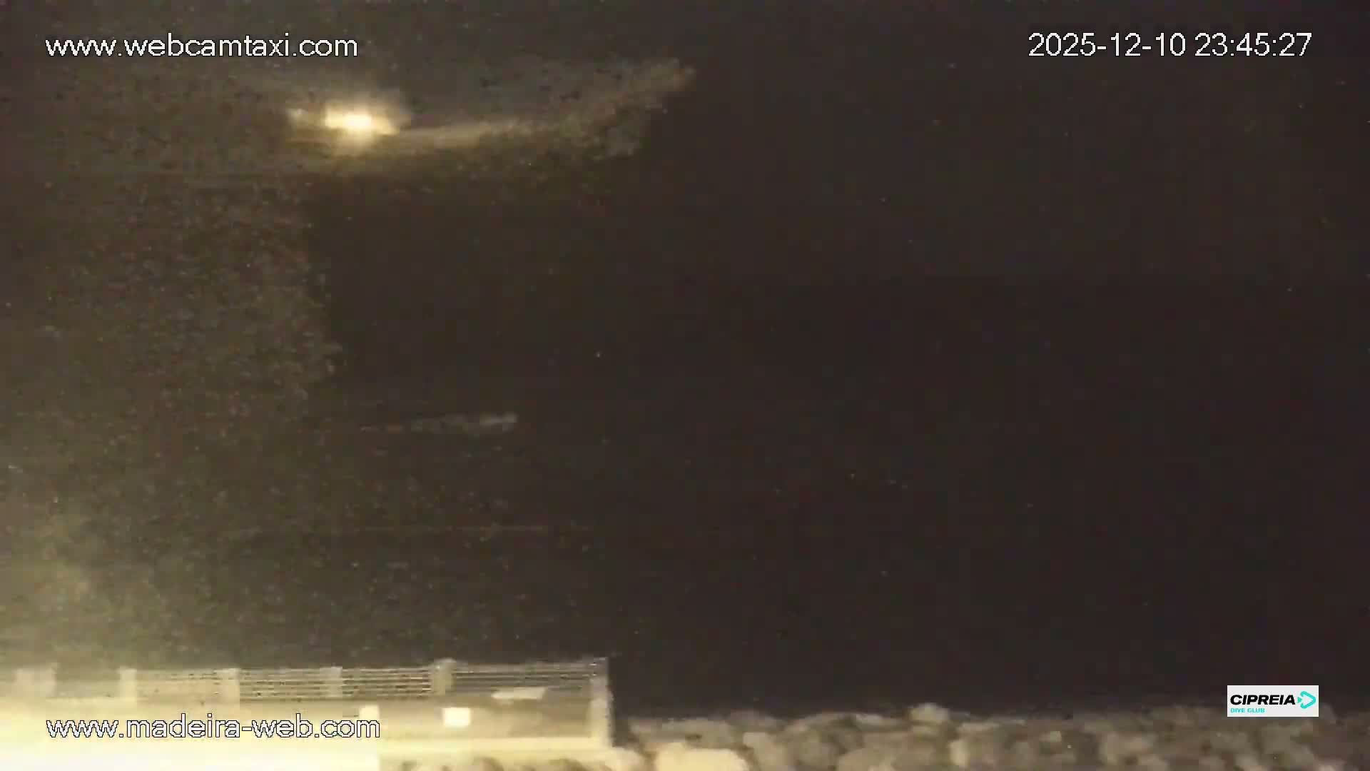 A blurry, dark outdoor scene at night shows a railing or structure in the foreground, with countless illuminated specks filling the air, indicative of heavy rain or mist.