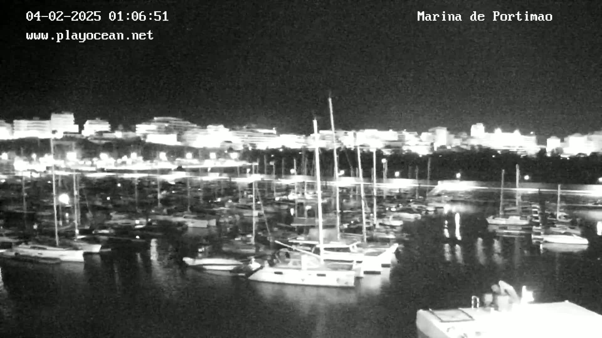 A nighttime, low-light view of a marina filled with boats, with a brightly lit city in the background.