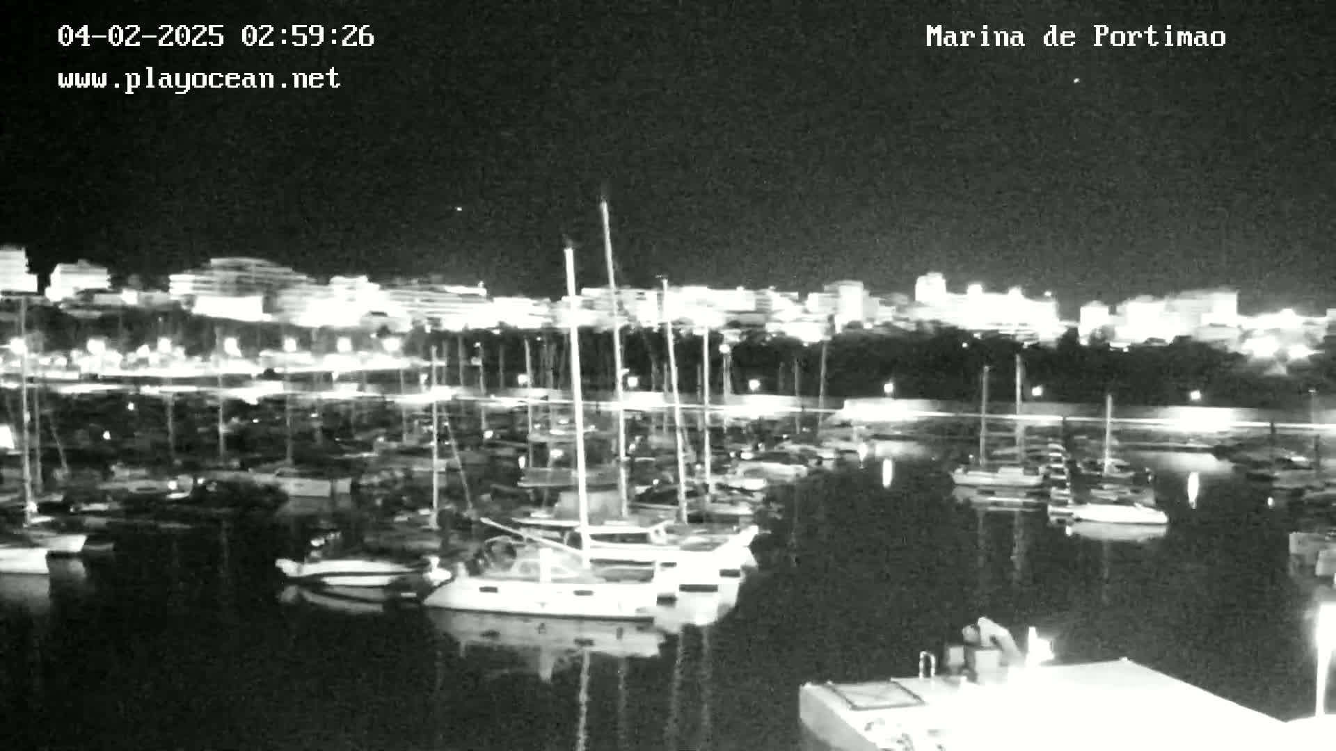 At night, a marina filled with boats is illuminated by city lights across the water.