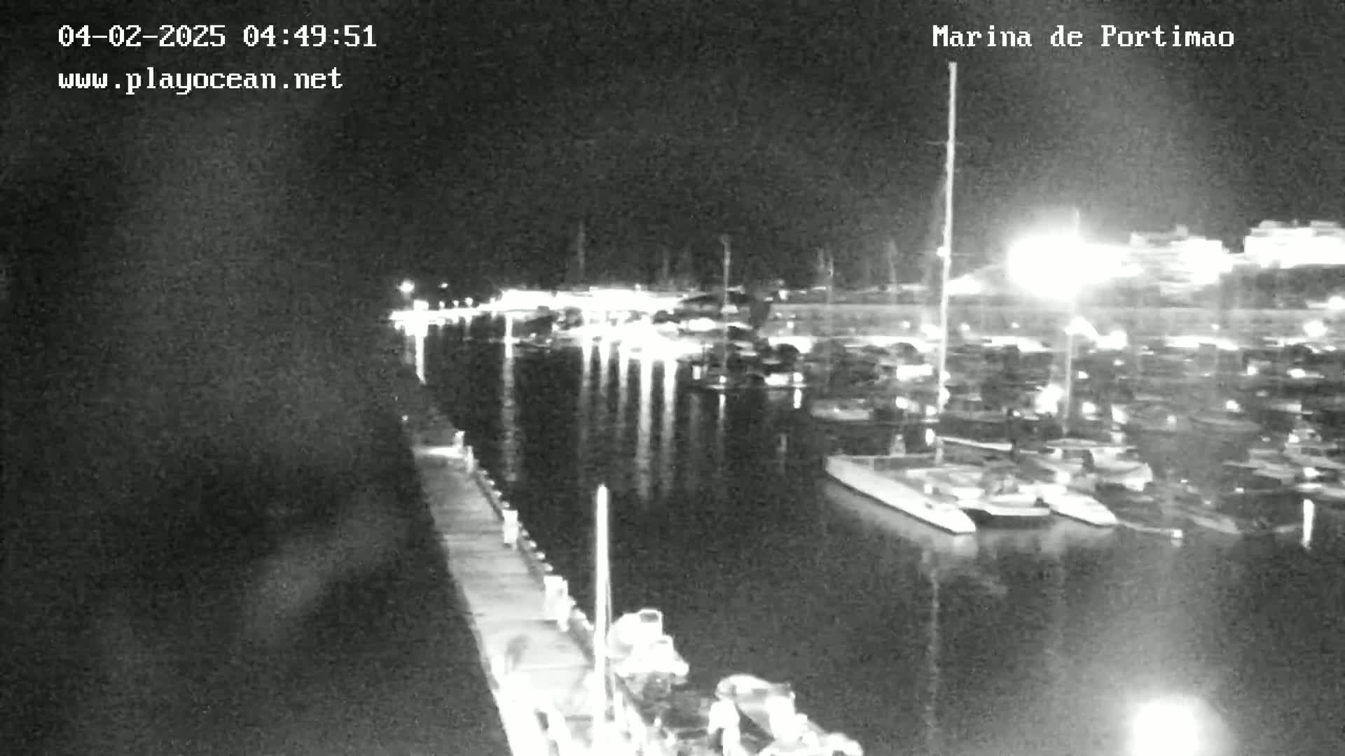 A nighttime, black and white view of a marina filled with boats, calm water reflecting lights from the docks and surrounding buildings.