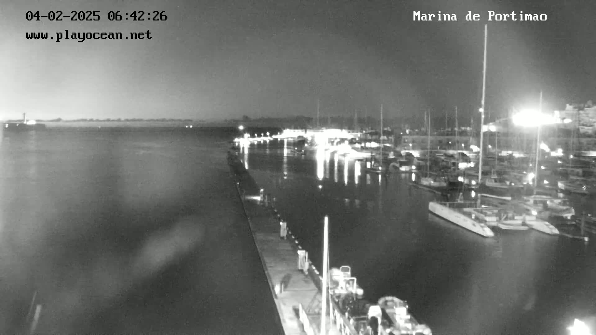 A nighttime, low-light view of a marina filled with boats, calm water reflecting the lights, and a pier extending into the scene.