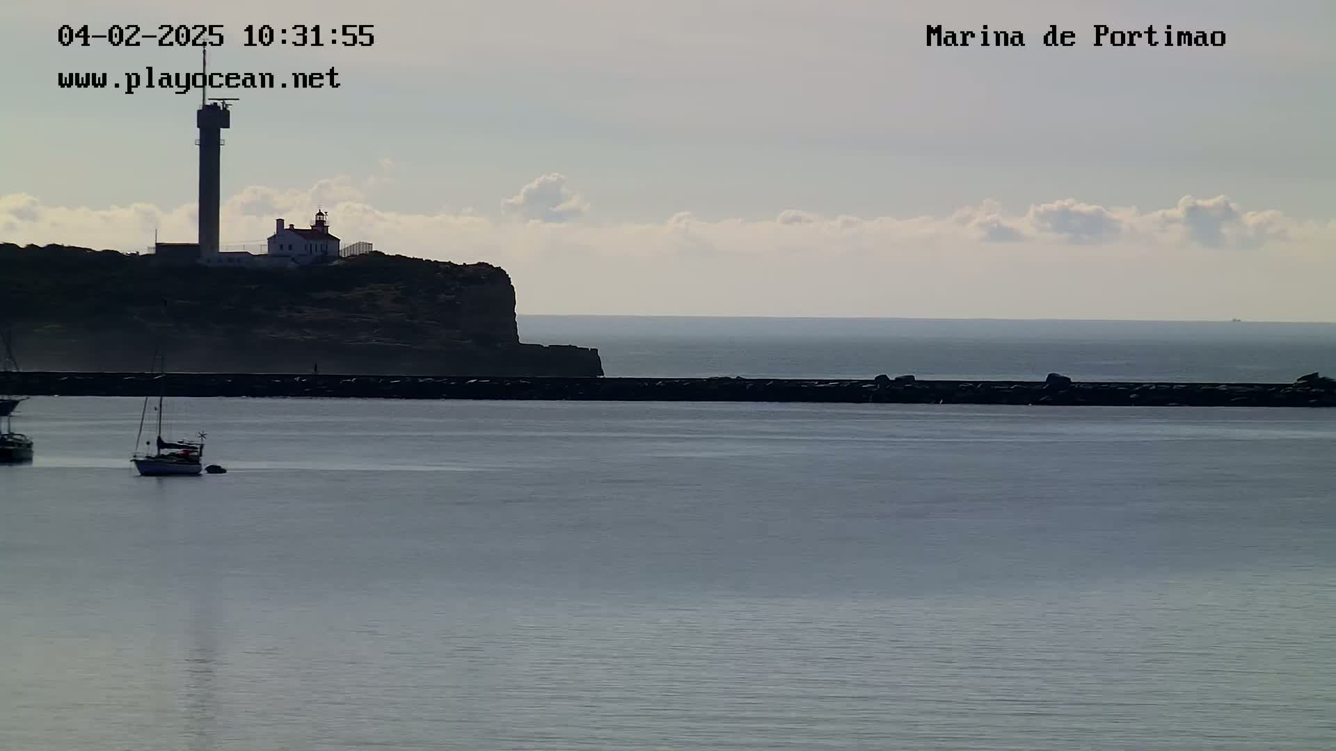 A lighthouse sits atop a dark cliff overlooking a calm body of water with a few sailboats and a breakwater.