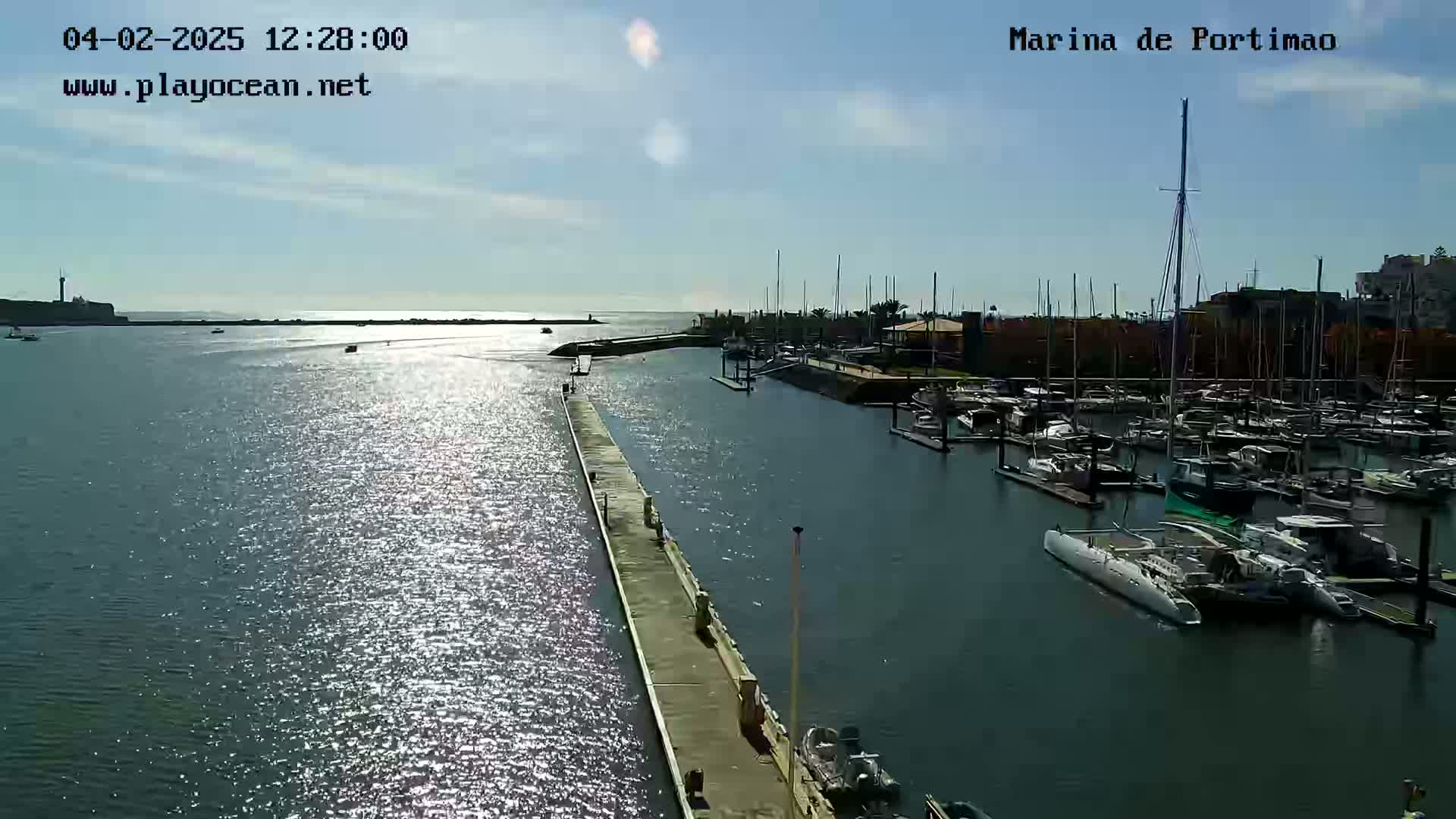 A marina filled with numerous boats is seen on a sunny day, with calm water reflecting sunlight.