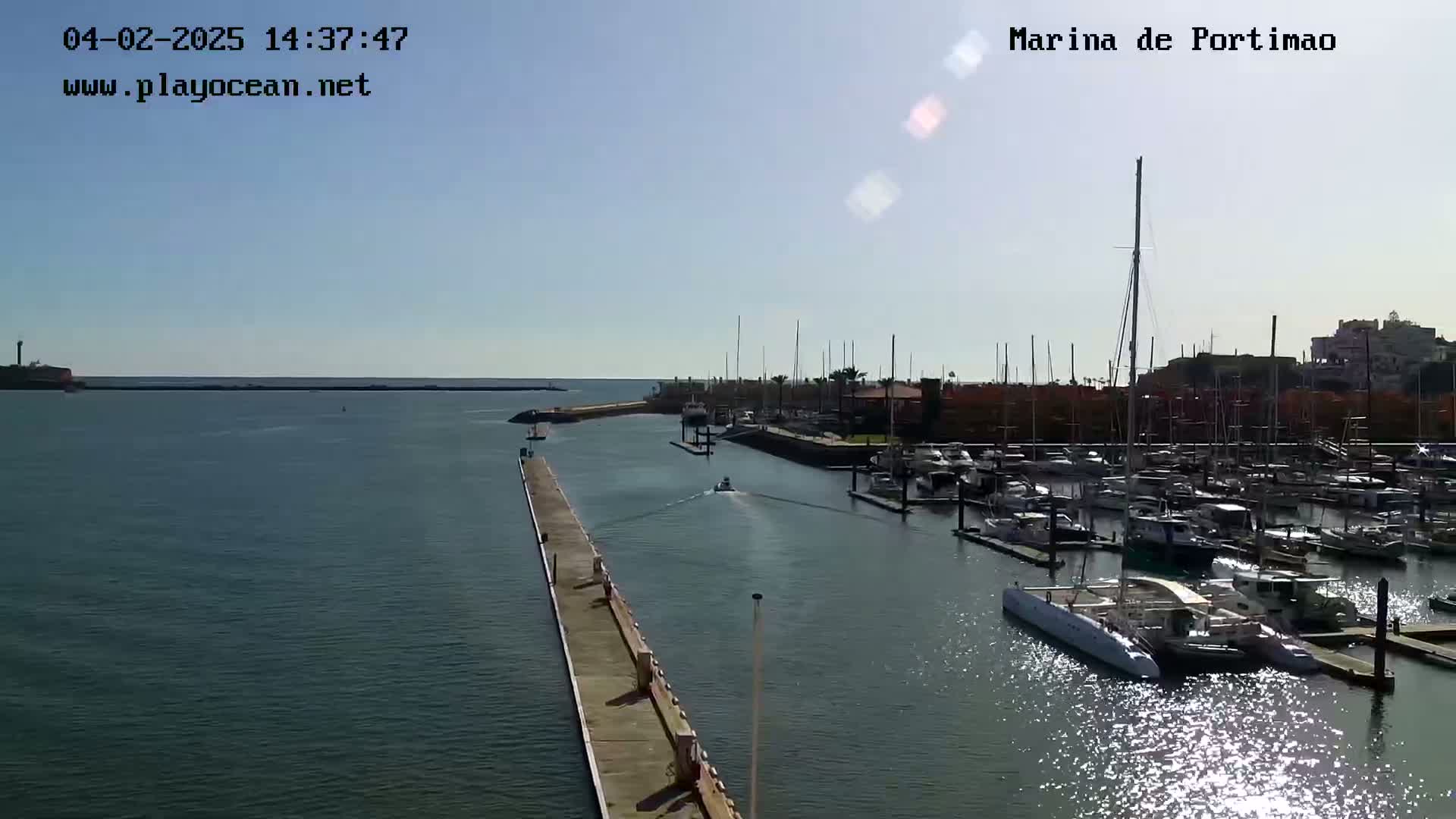 A sunny day shows a marina filled with boats and yachts, a long pier extending into the calm water.