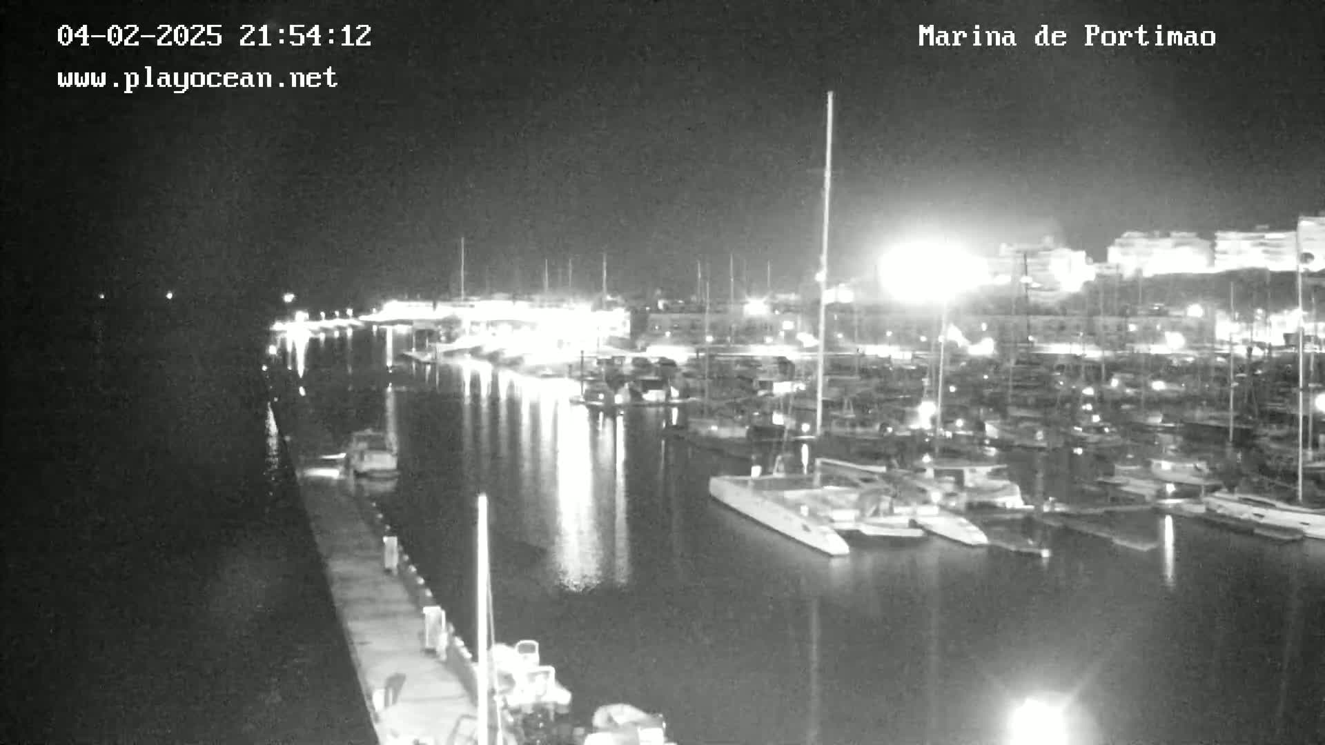 A nighttime, black and white view of a marina filled with many boats and yachts, illuminated by lights along the docks and buildings in the background.