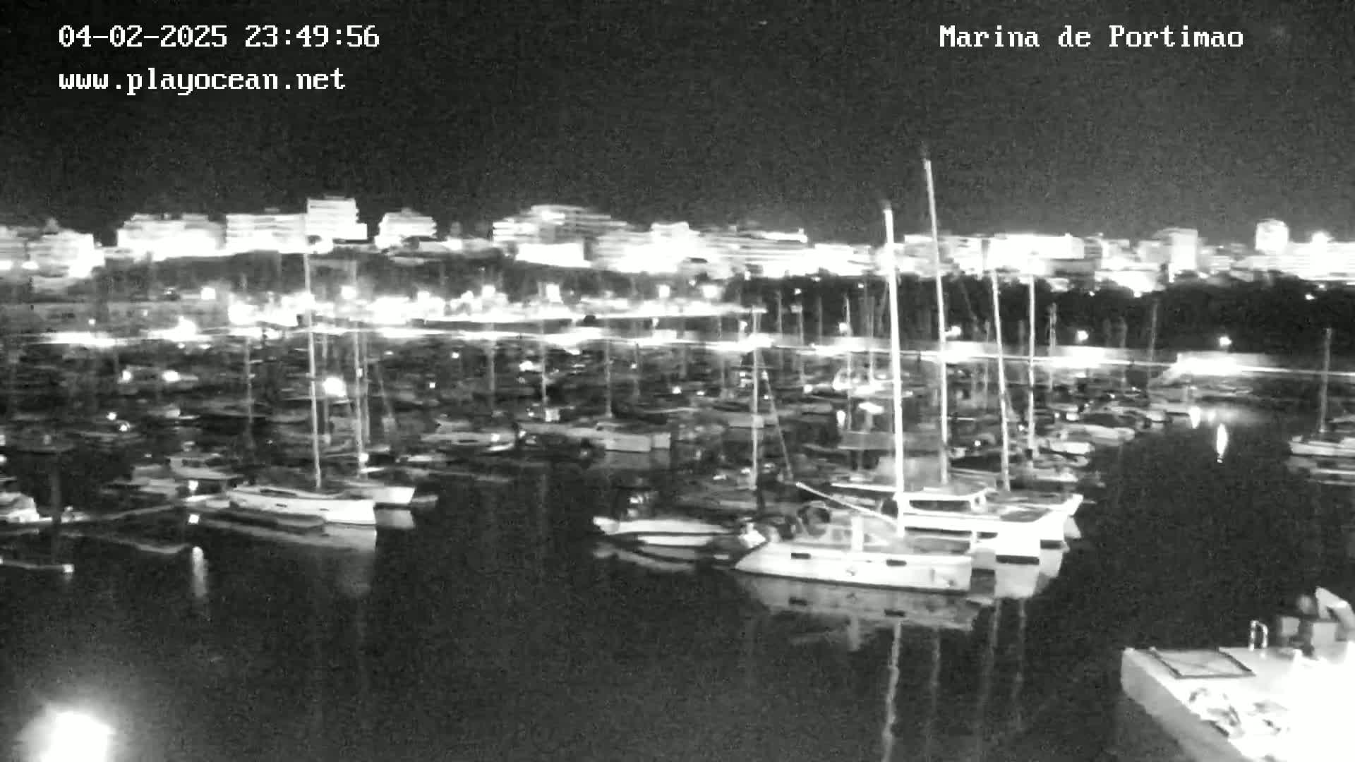 A nighttime view of a marina filled with numerous boats and yachts, illuminated by lights reflecting on calm water, with a city skyline visible in the background.