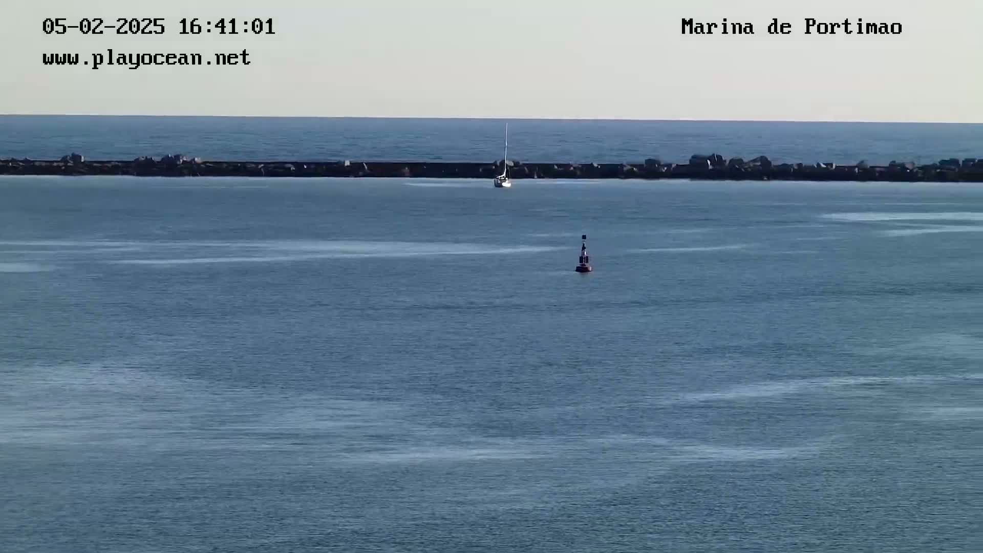 A sailboat and a buoy are visible in calm waters beyond a long, low breakwater under a clear sky.