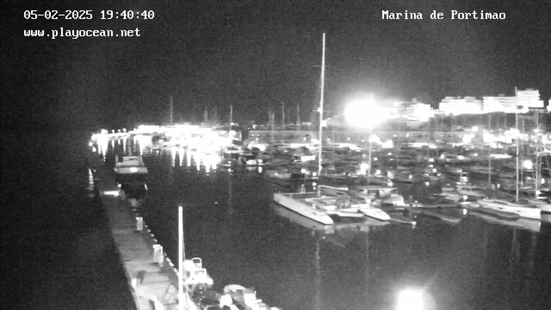 A nighttime, black-and-white view of a marina filled with numerous boats and yachts moored along a dock.