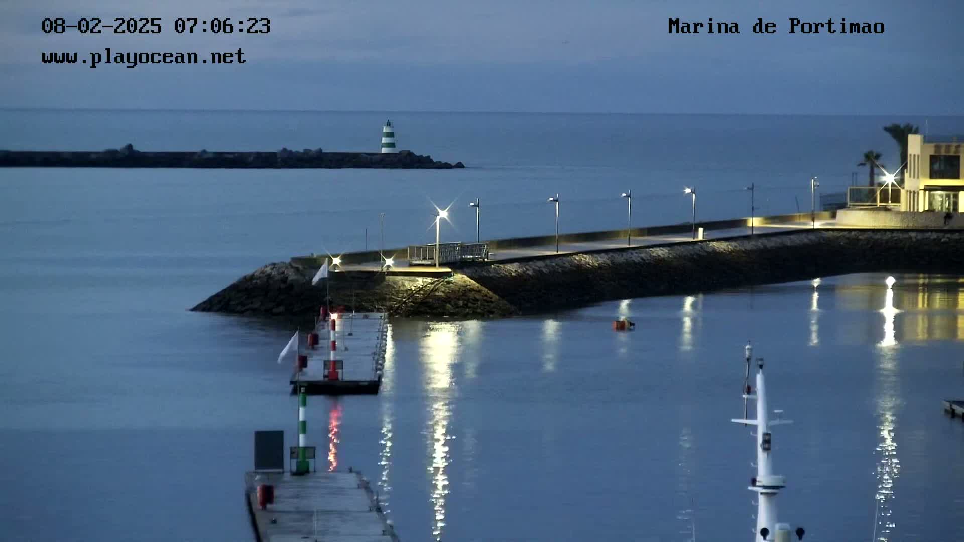A calm, dark-blue harbor at twilight is lit by artificial lights along a seawall, with a lighthouse visible in the distance.