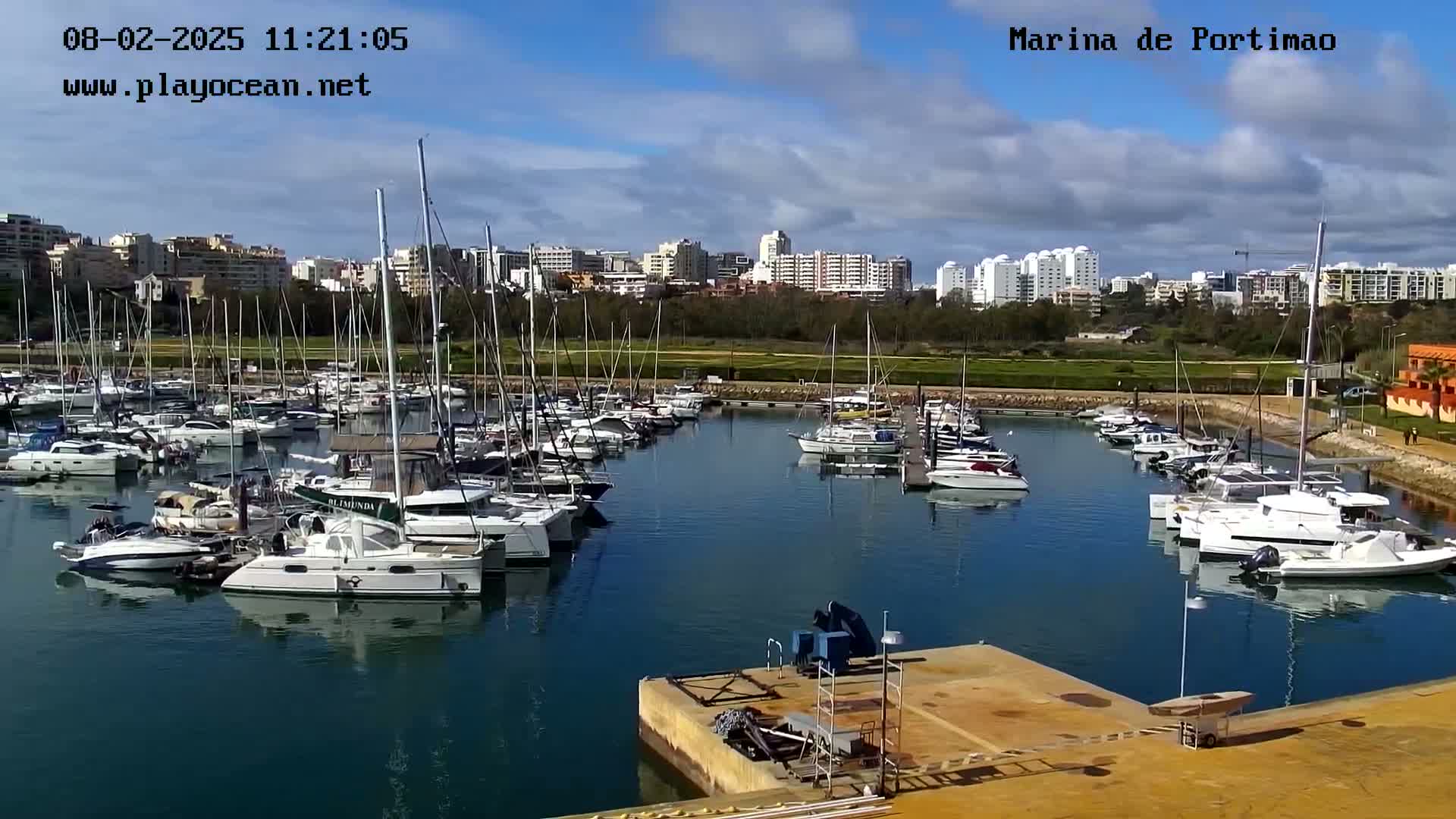 A marina filled with numerous sailboats and motorboats is situated in front of a city skyline under a partly cloudy sky.