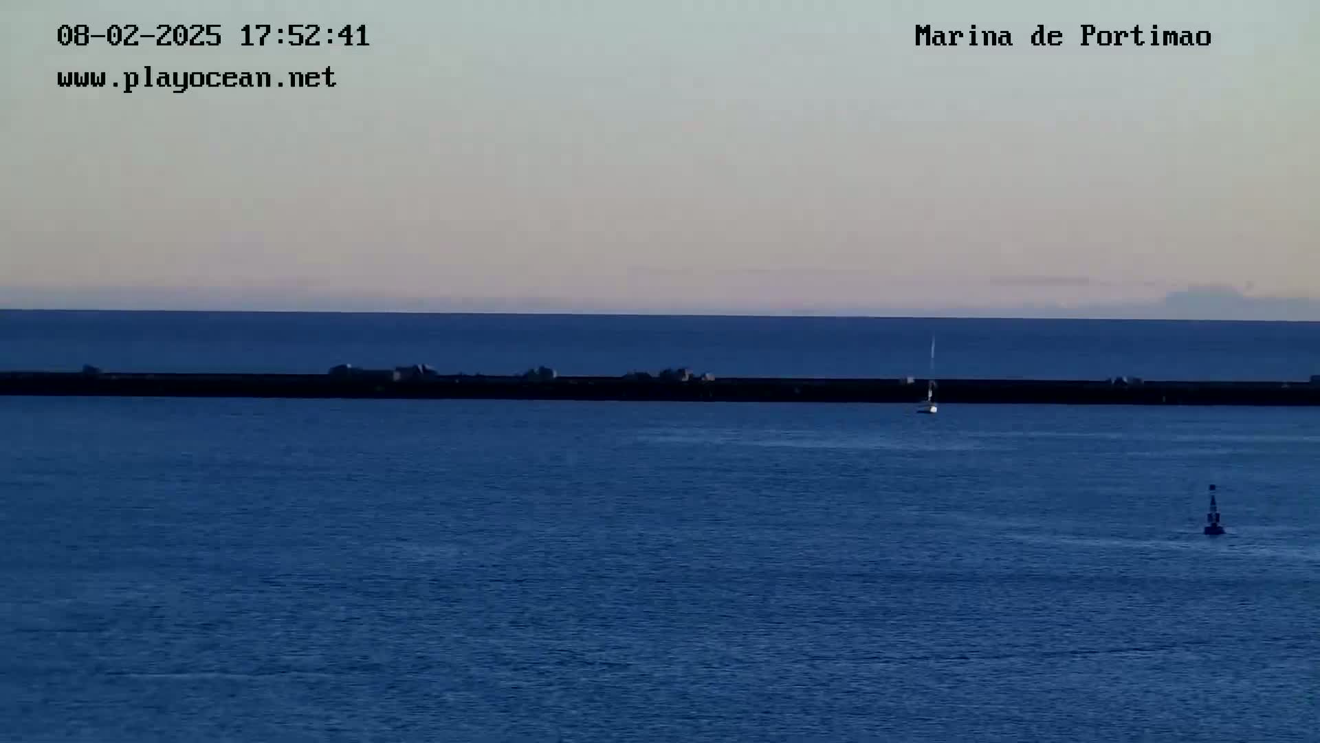 A calm, dusky-blue ocean stretches to the horizon, featuring a long, dark breakwater with a lone sailboat and a small marker buoy in the water.