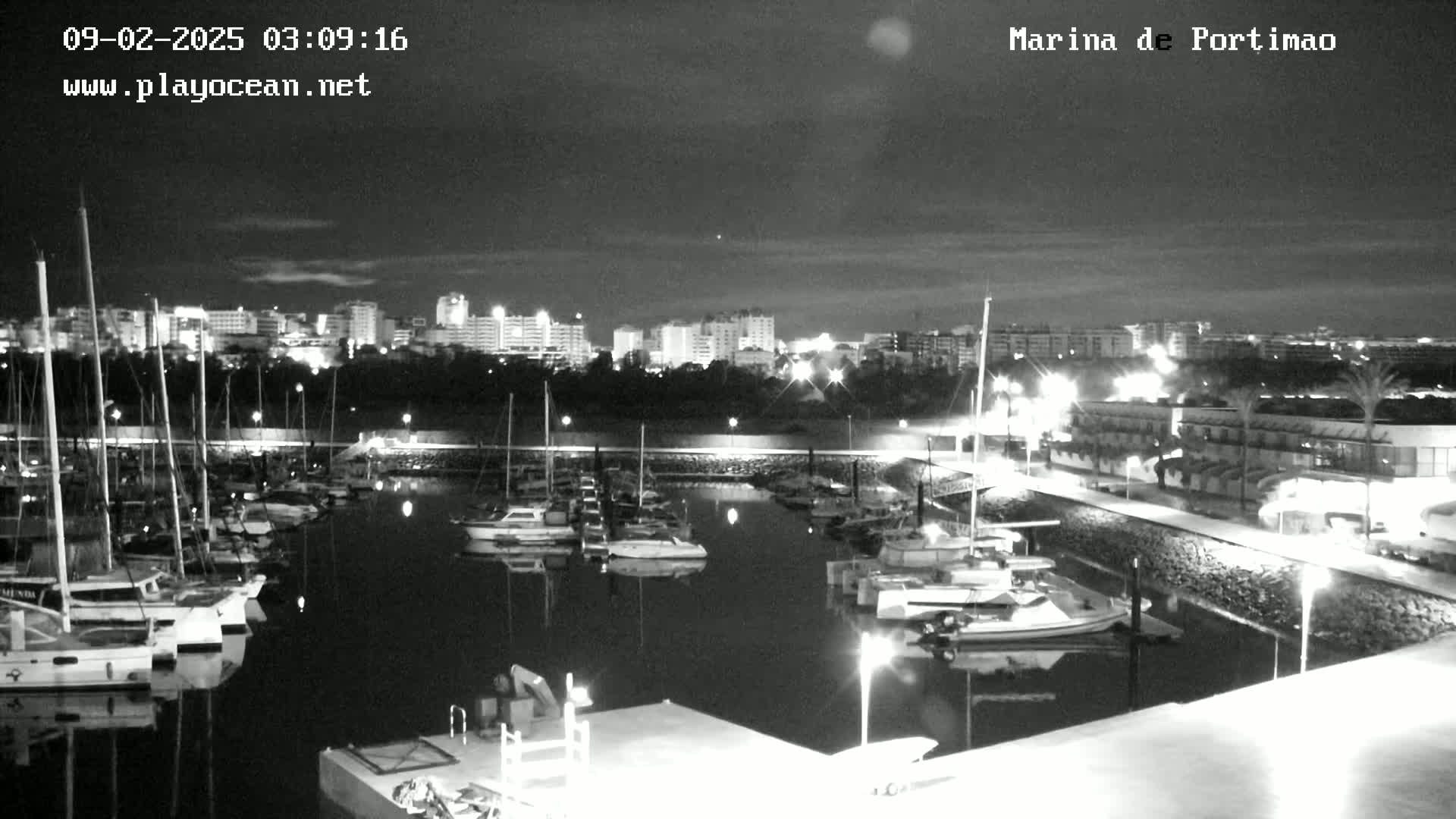 A nighttime, black and white view of a marina filled with boats,  with a city skyline visible in the background under a partly cloudy sky.