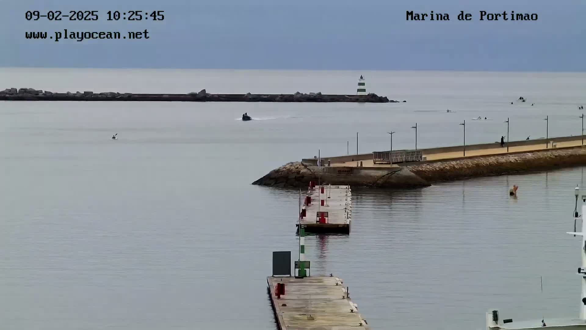 A calm, overcast day shows a marina with a long dock extending into the water, a small jetty, a lighthouse in the distance, and several small watercraft.