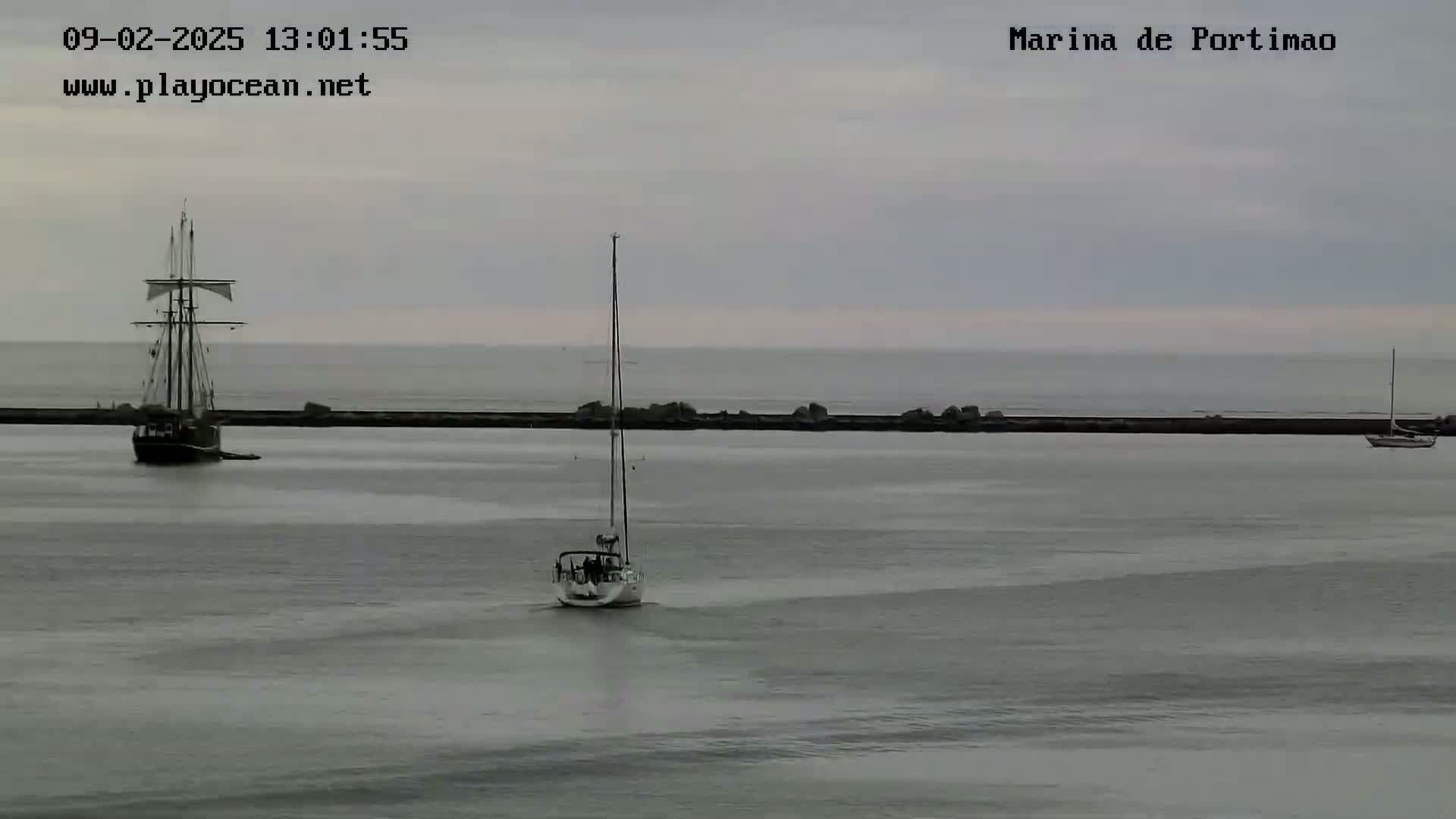 A sailboat and a large sailing ship are moored in calm, overcast waters behind a low breakwater.