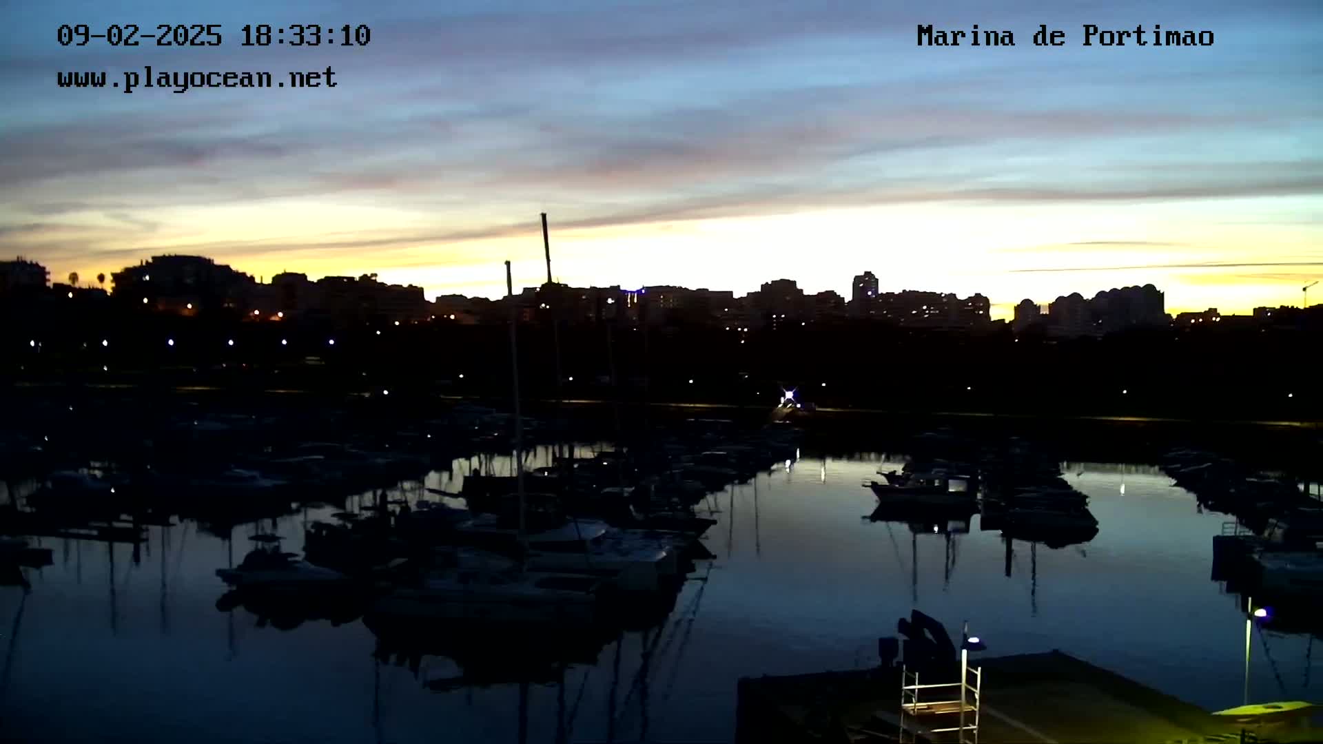 Many boats are moored in a marina at dusk, under a partly cloudy sky with the last light of sunset.