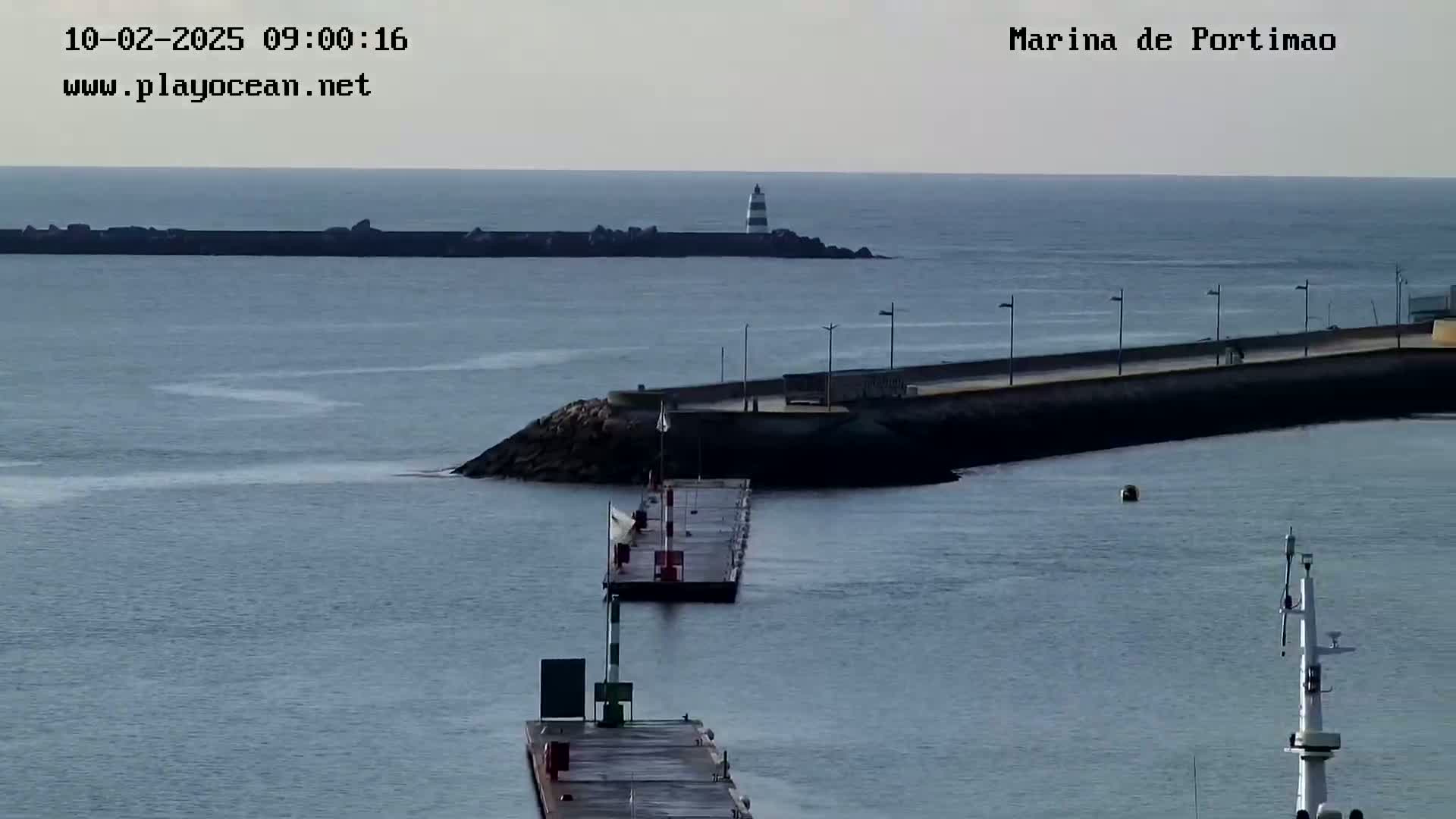 A calm, gray day shows a harbor with a breakwater, a lighthouse in the distance, and a floating dock.
