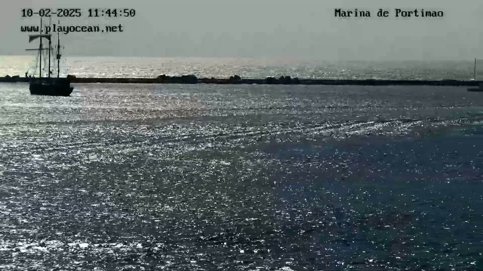 A three-masted sailing ship is moored near a breakwater in a calm, sunlit body of water.