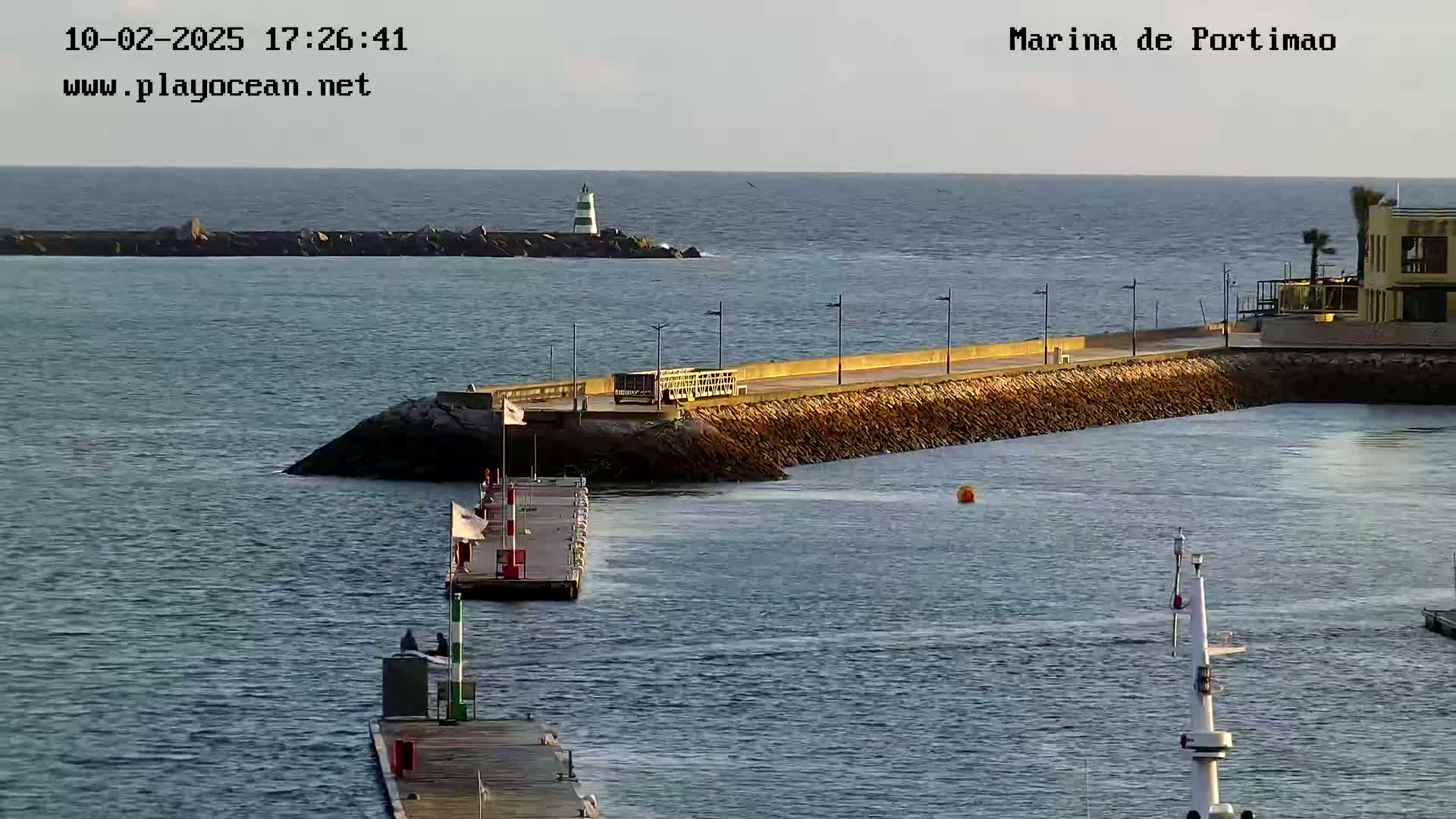 A calm, sunny day at a marina shows a harbor wall, a small dock, and a lighthouse in the distance.