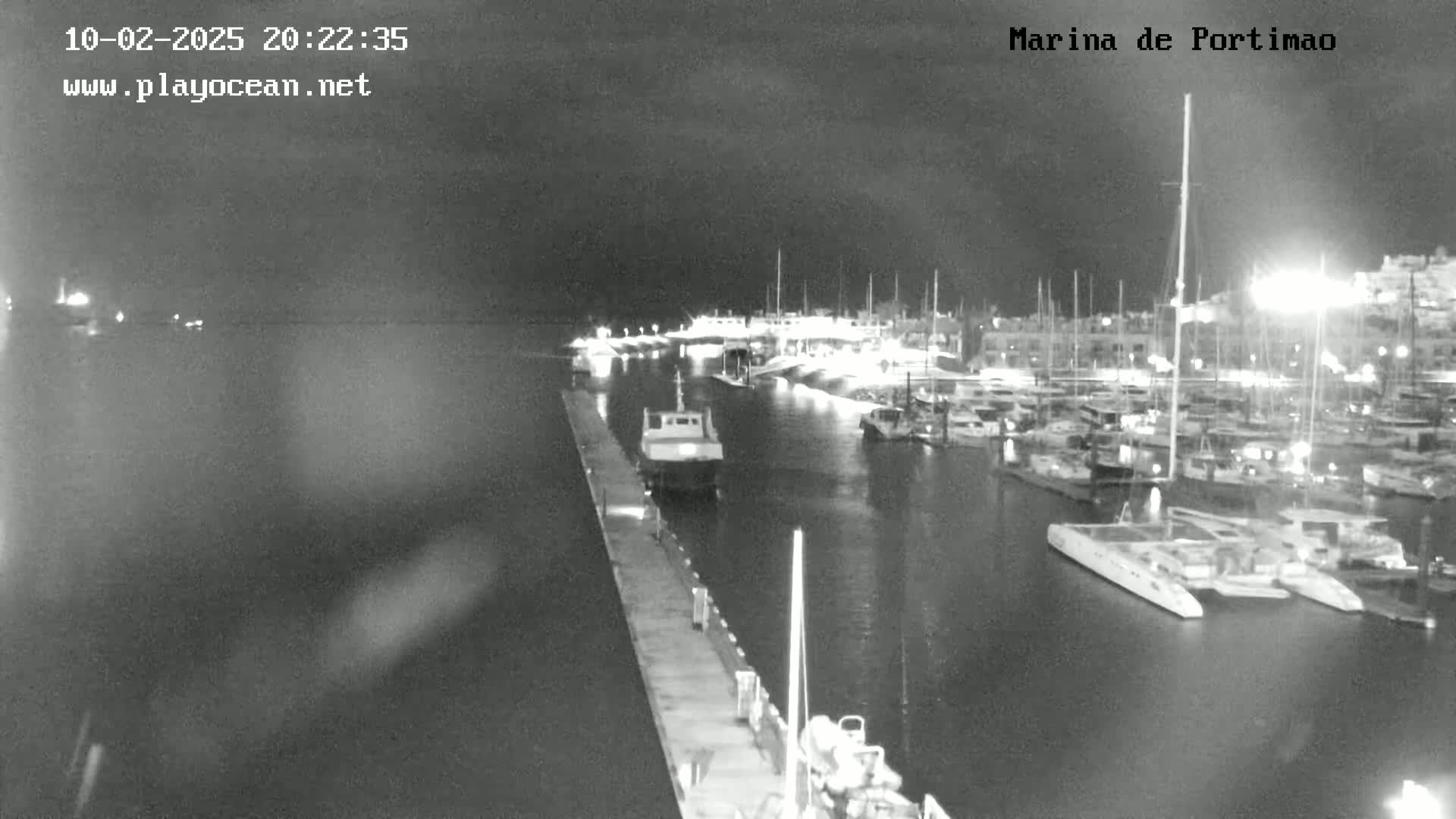 A nighttime, black and white view of a marina filled with docked boats and yachts, illuminated by lights along the docks and on the boats themselves.
