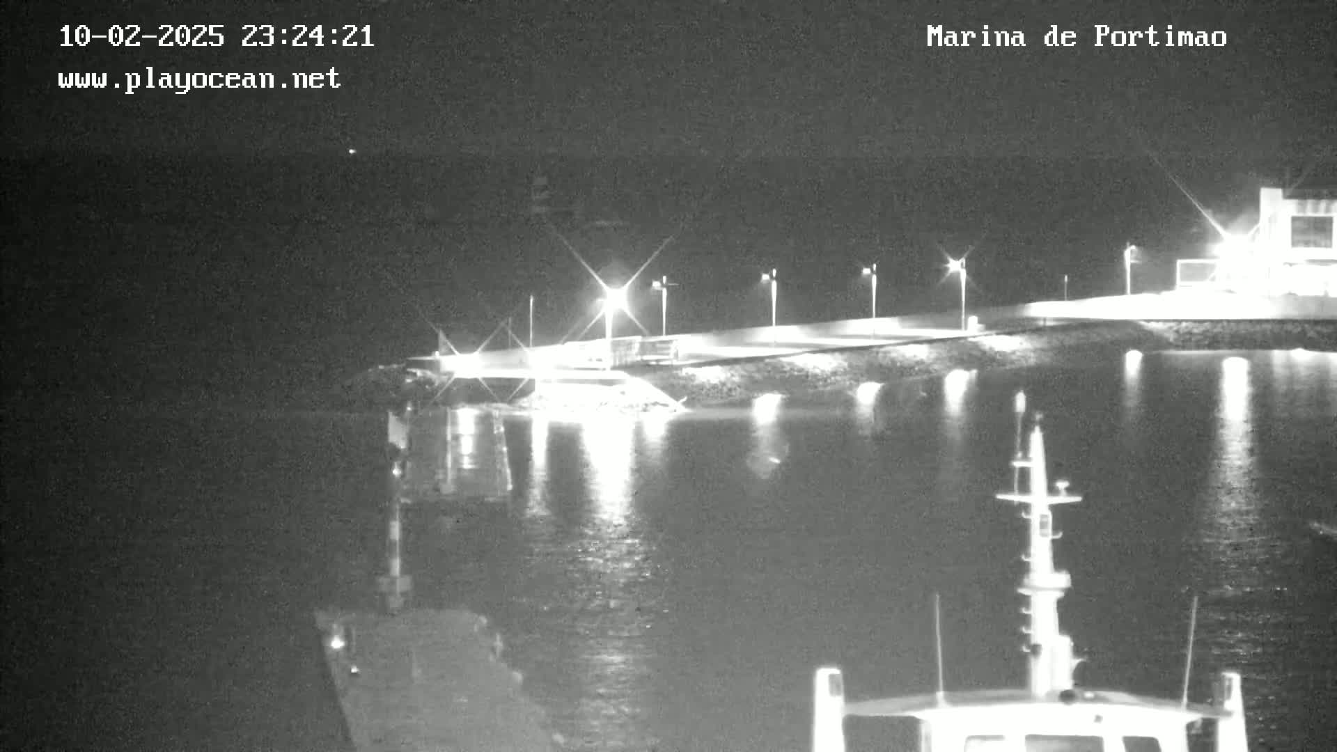 A nighttime, low-light view of a marina, with calm water reflecting lights from the shore and a boat in the foreground.