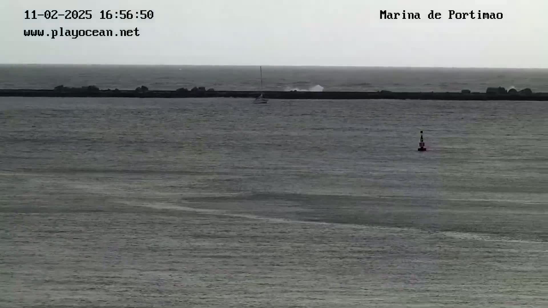 A sailboat is visible in the calm waters beyond a wave-breaking barrier on an overcast day.