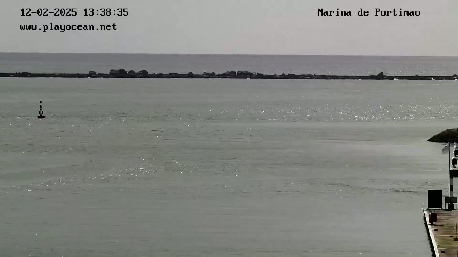 A calm, gray body of water is seen with a long, low rock barrier extending across the middle ground, and a small portion of a dock visible in the bottom right corner.