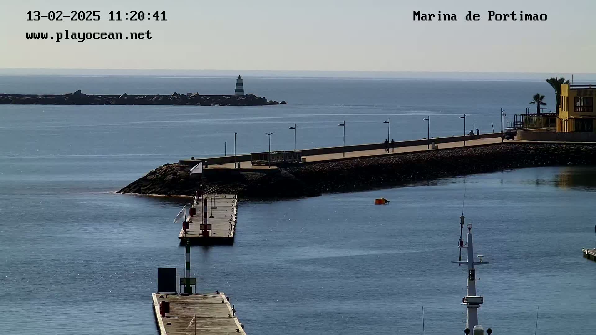 A marina with calm, blue water, a lighthouse in the distance, and a paved walkway along a breakwater on a sunny day.