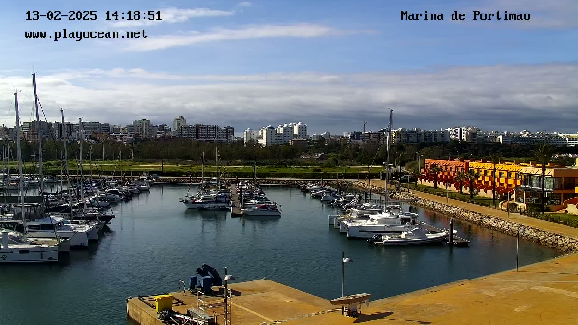 A marina filled with sailboats and motorboats is situated next to a waterfront building under a partly cloudy sky.
