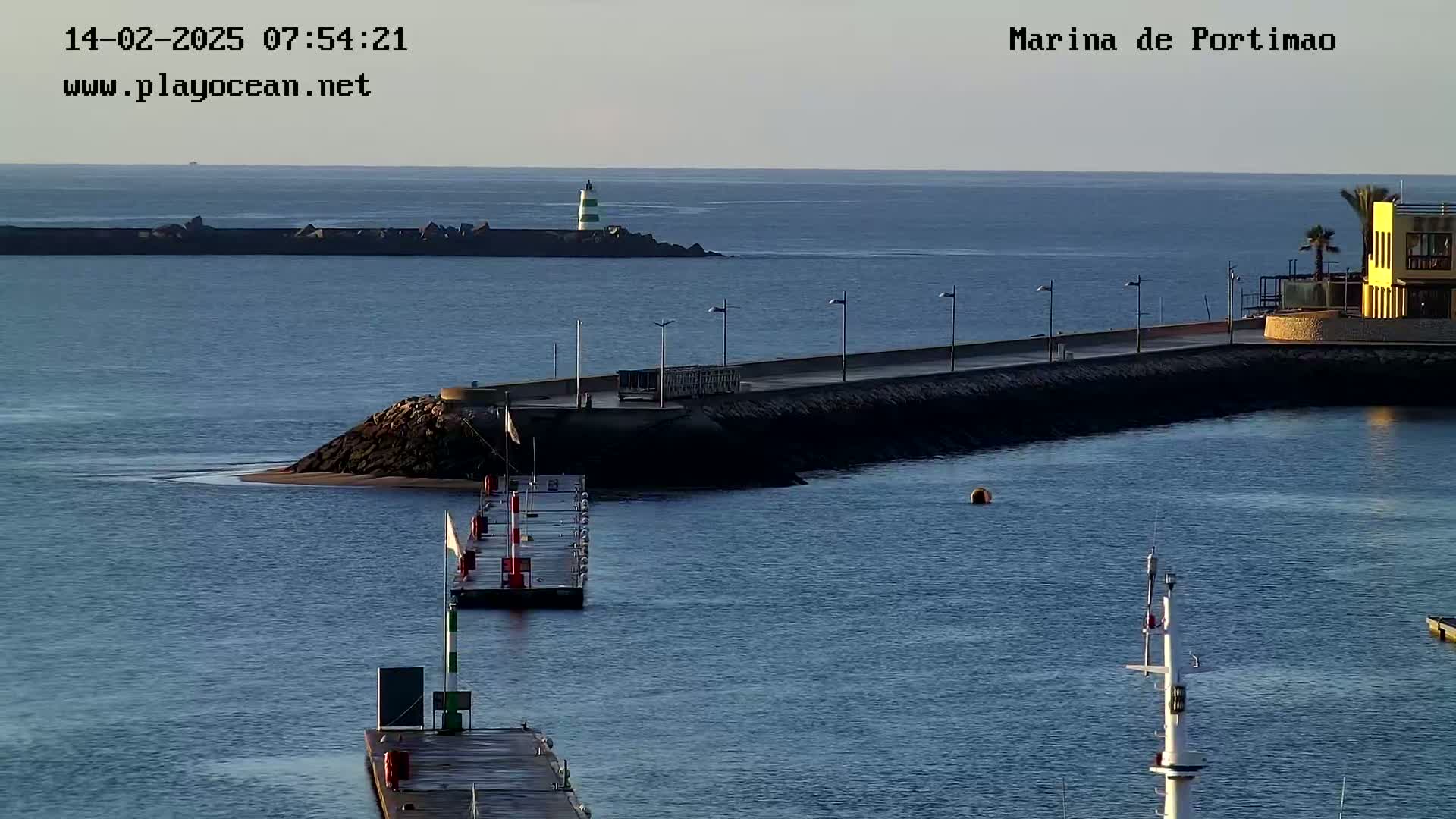 A calm harbor scene shows a breakwater, a lighthouse in the distance, a small dock, and a building on the shore under a clear sky.