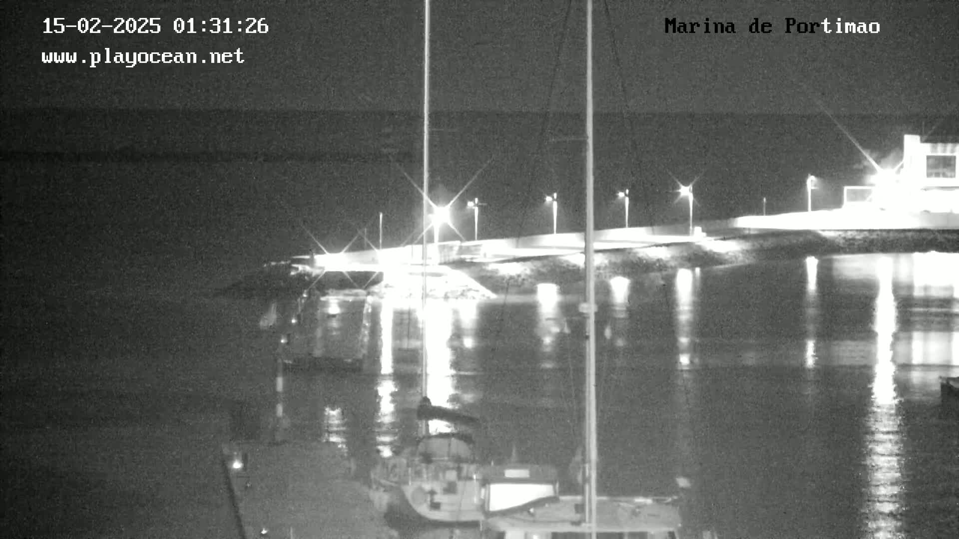 A nighttime, low-light view of a marina with boats moored, illuminated by lights along a nearby walkway.