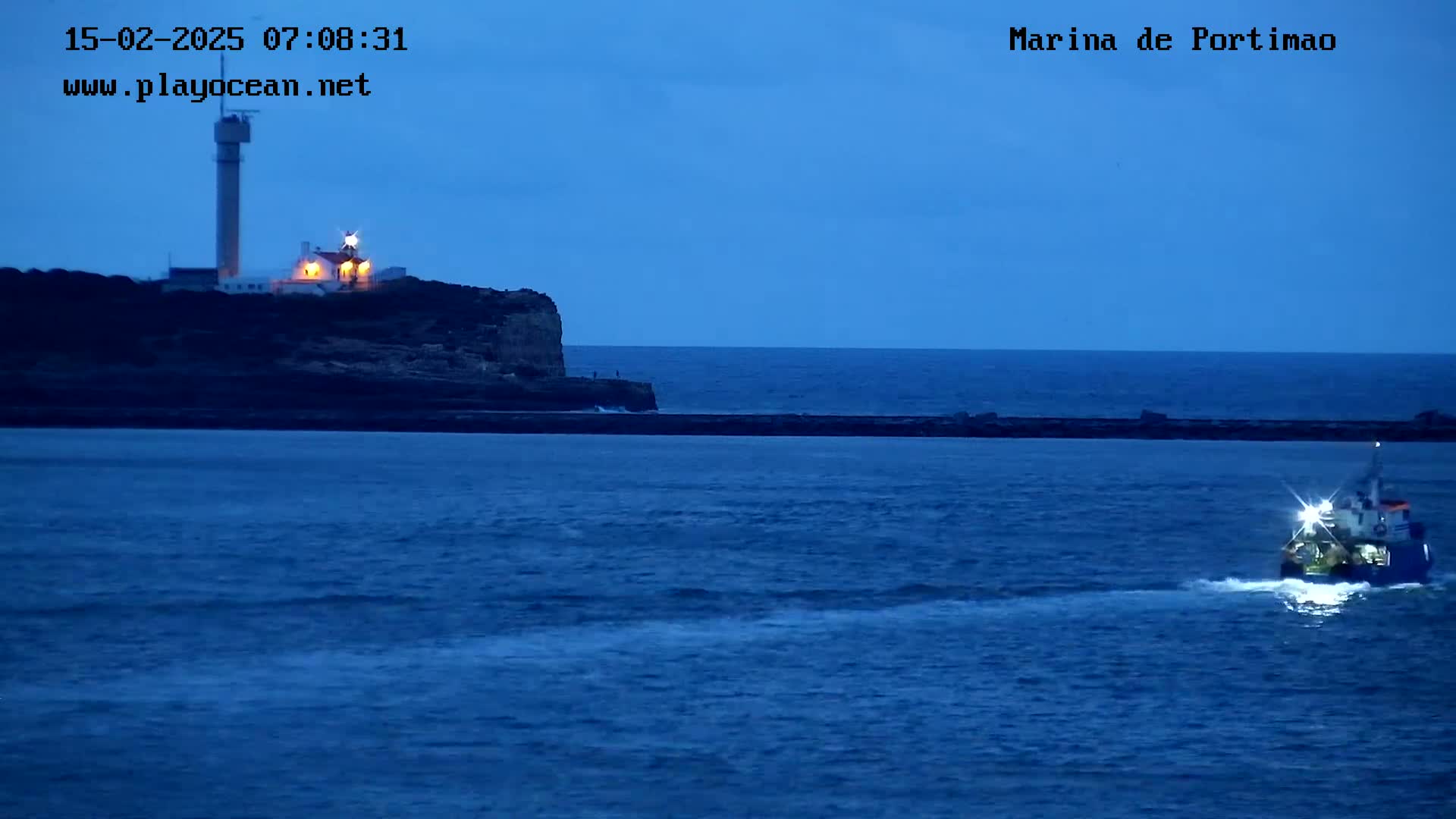 A small boat sails past a breakwater toward a lit lighthouse on a cliff at twilight.