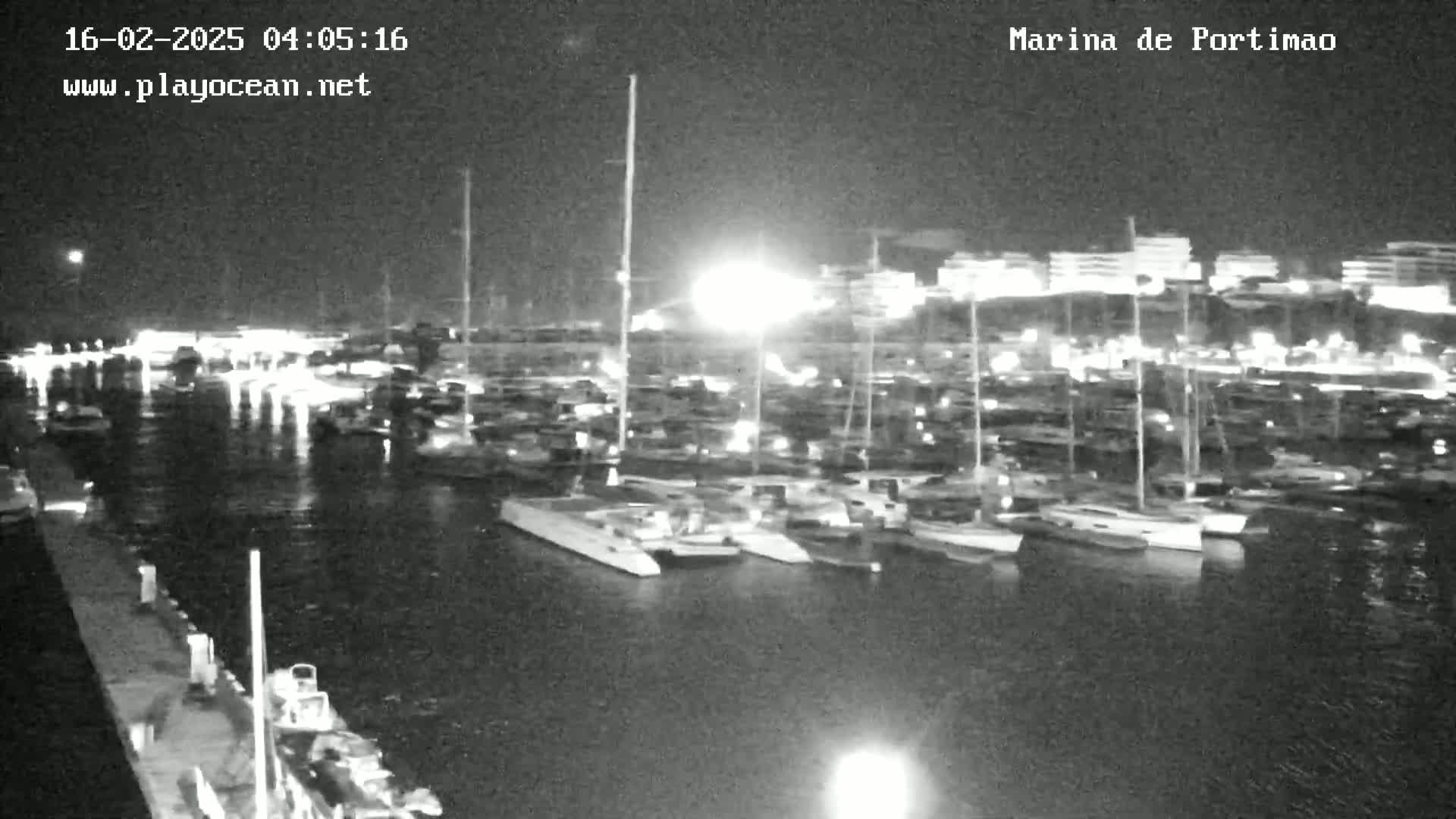 A nighttime, low-light view of a marina filled with numerous boats and yachts, docked alongside a waterfront.
