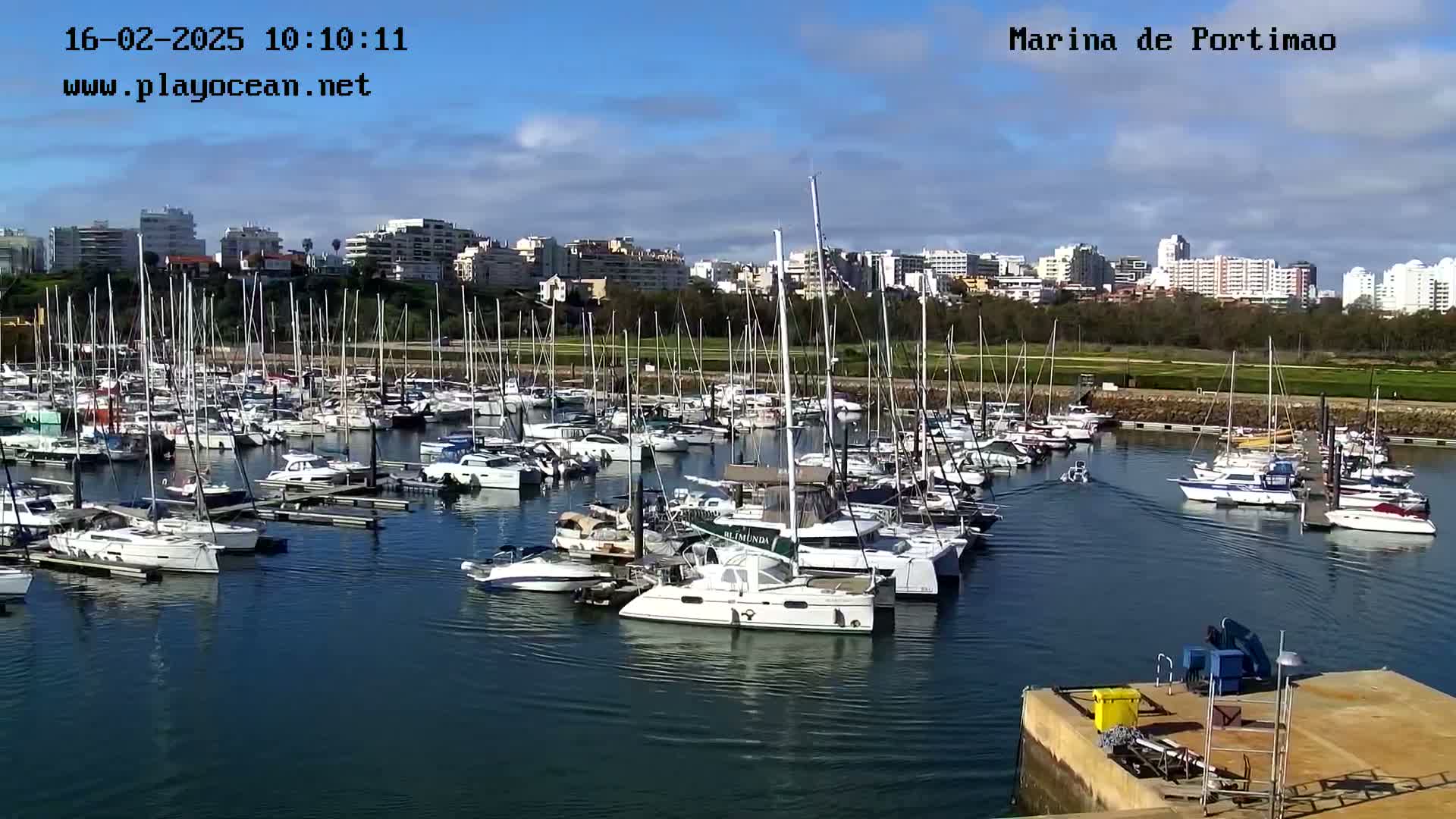 A marina filled with numerous sailboats and motorboats is situated in front of a city skyline under a partly cloudy sky.