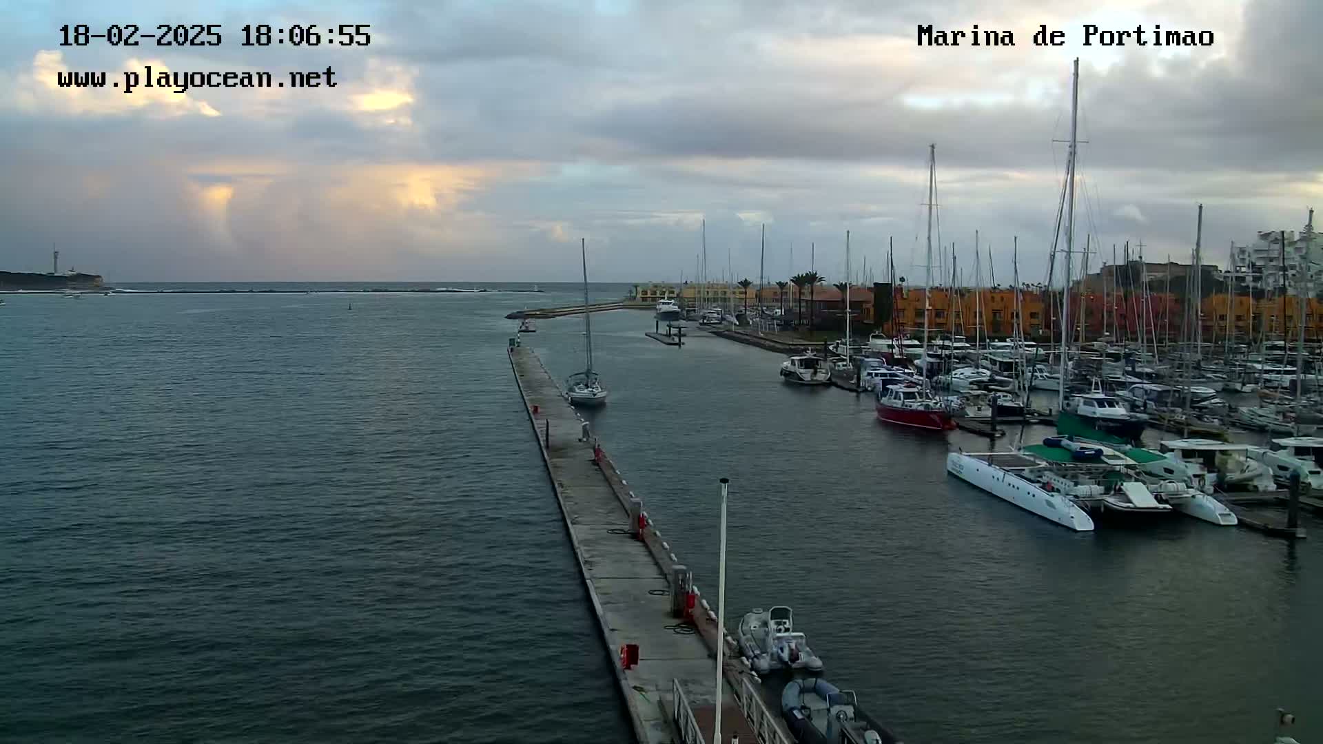 A marina filled with numerous boats and yachts is seen under a cloudy sky, with a calm sea extending to a distant coastline.