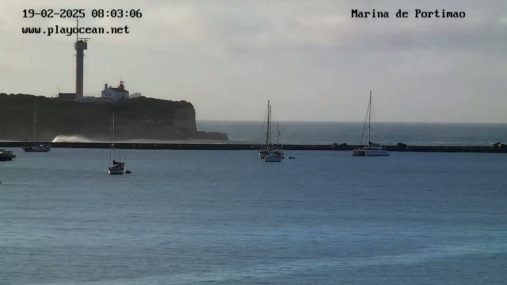 Several sailboats are moored in a calm harbor next to a rocky cliff with a lighthouse and tower under an overcast sky.