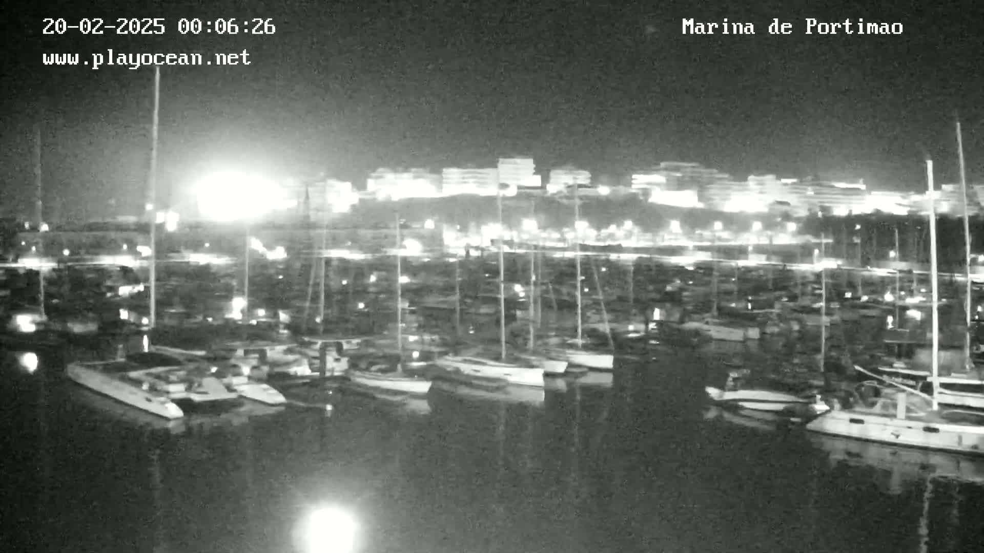 A nighttime, low-light view of a marina filled with numerous boats docked alongside a brightly lit shoreline.