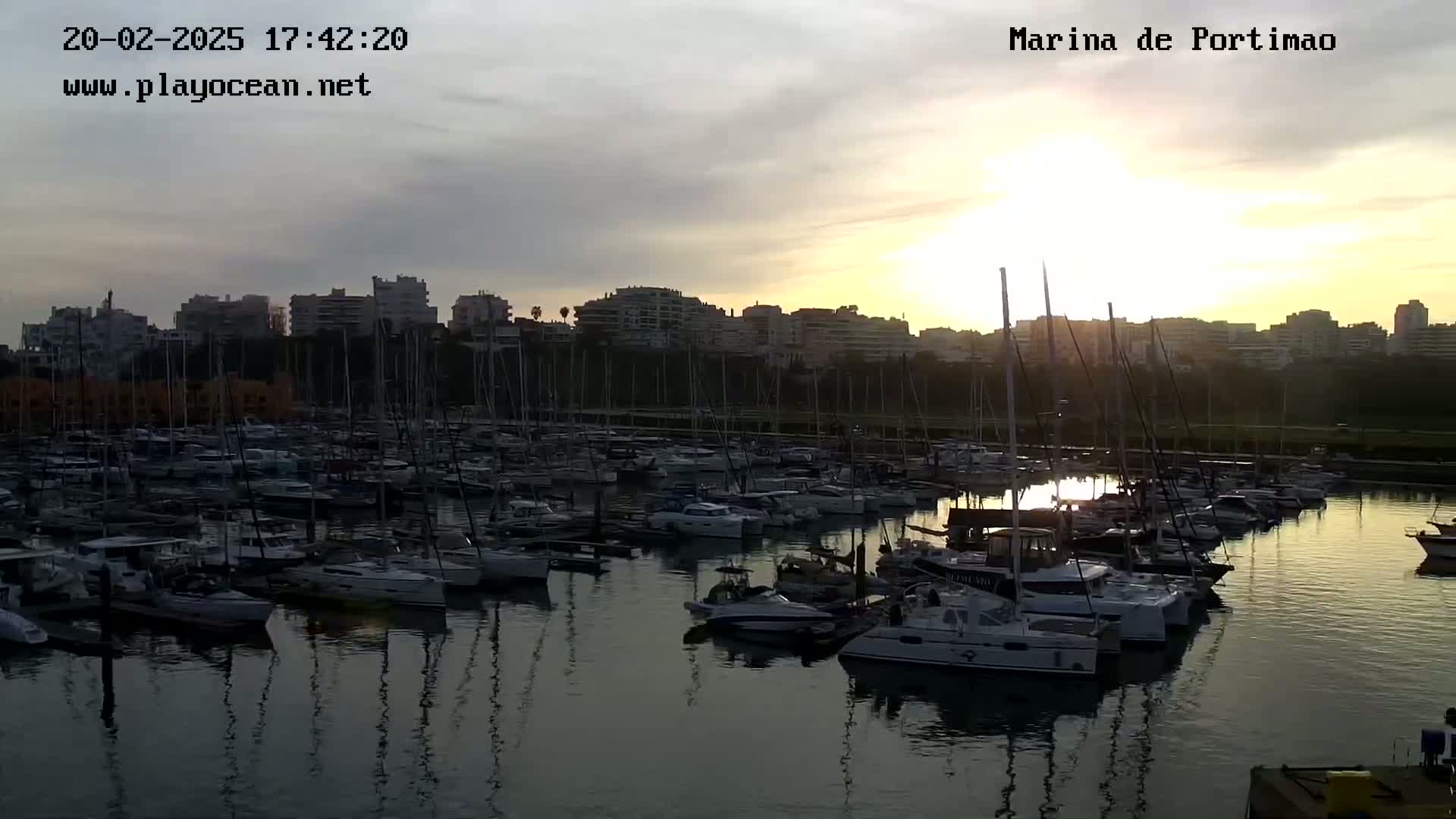 Many sailboats are docked in a marina at sunset under a partly cloudy sky.