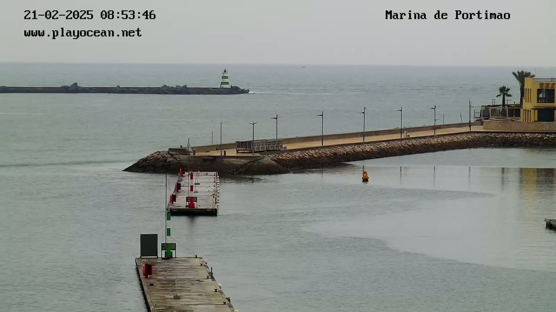 A marina with calm, gray water under an overcast sky, featuring docks, a breakwater, a lighthouse in the distance, and a building on the shore.