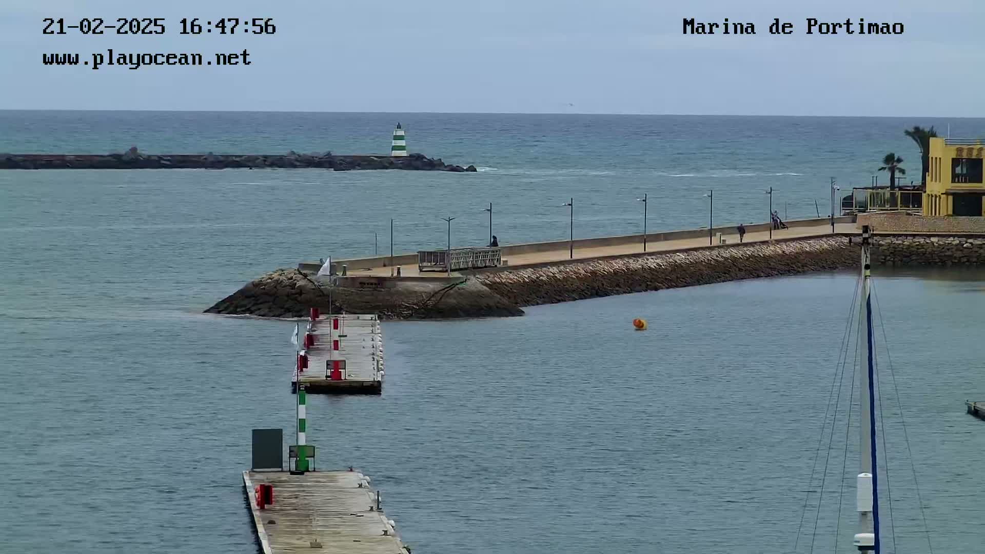 A harbor with calm water, a long paved walkway, a lighthouse, and a few people walking along the breakwater under an overcast sky.