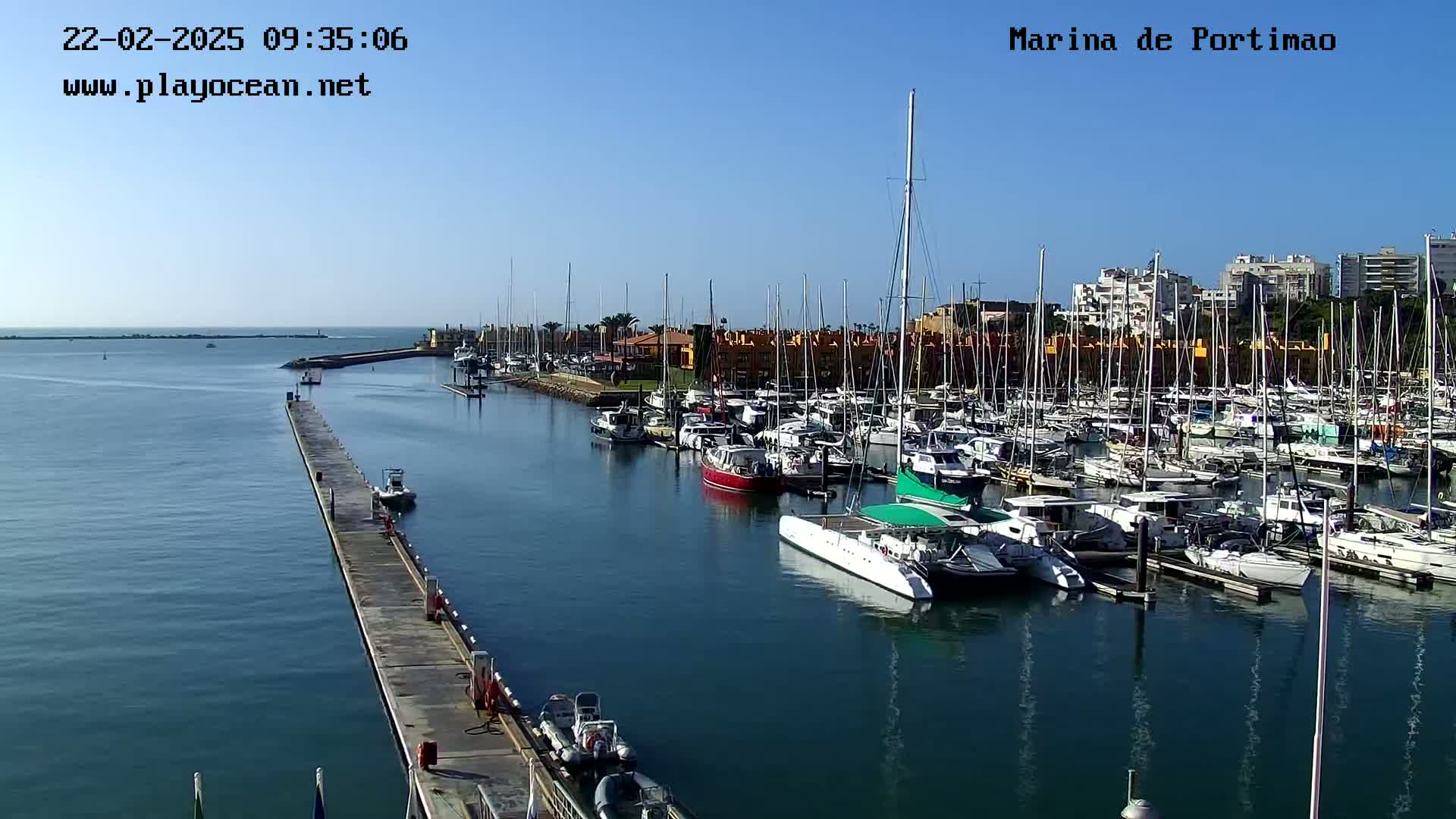 A marina filled with numerous sailboats and motorboats on a sunny day, with a long pier extending into the calm water.
