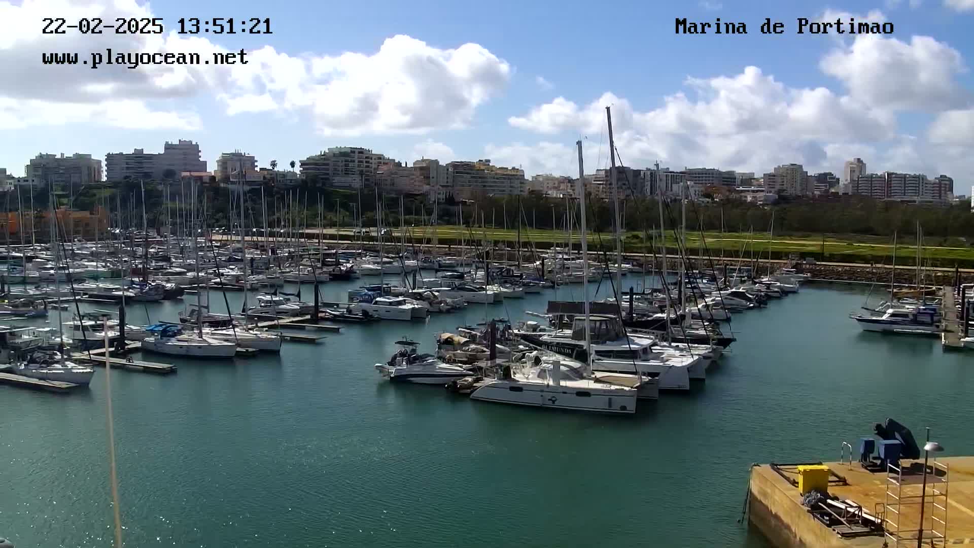 A marina filled with numerous sailboats and motorboats is seen under a partly cloudy sky.