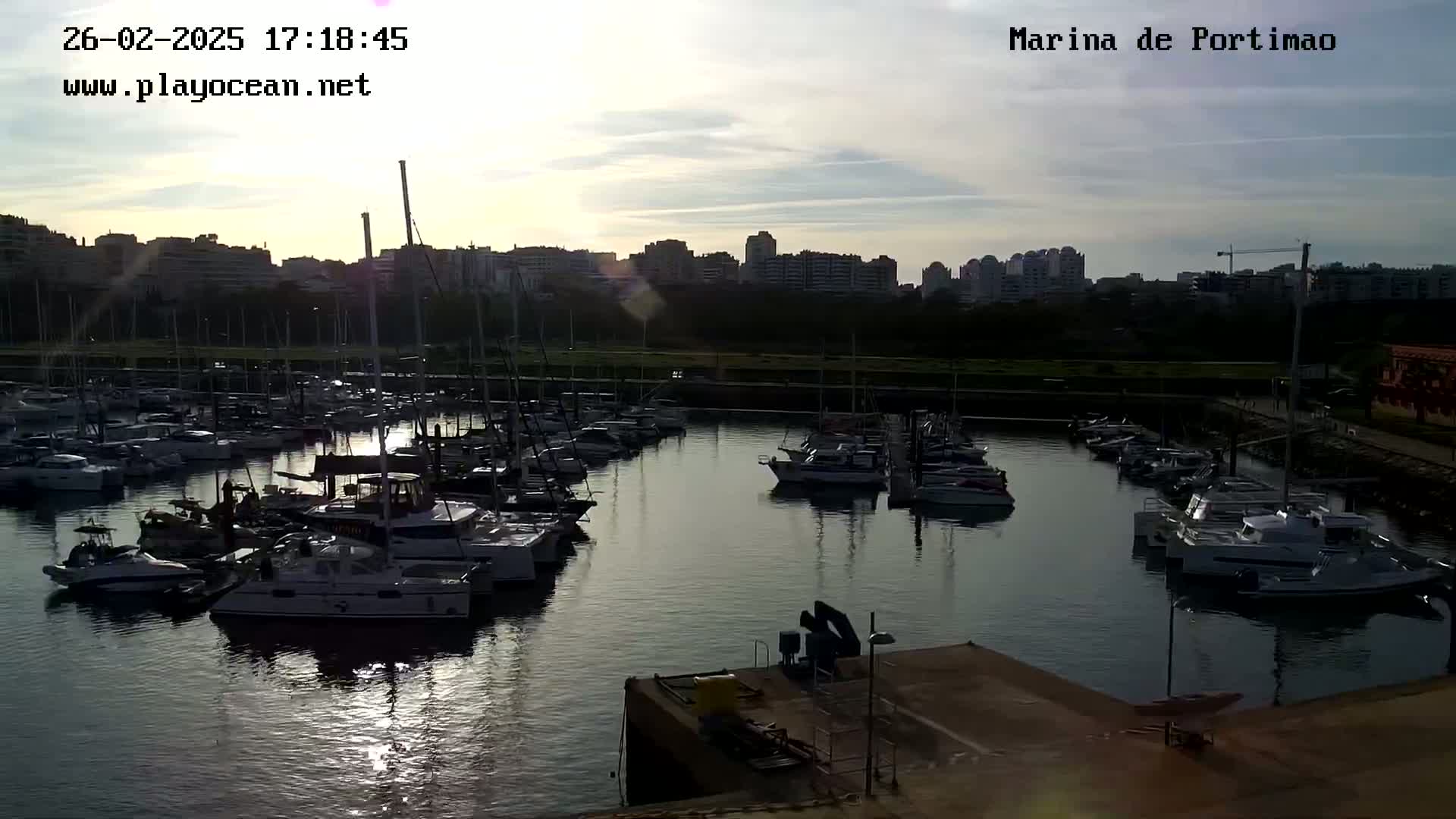 A marina filled with numerous boats is situated under a partly cloudy sky at sunset, with a cityscape visible in the background.