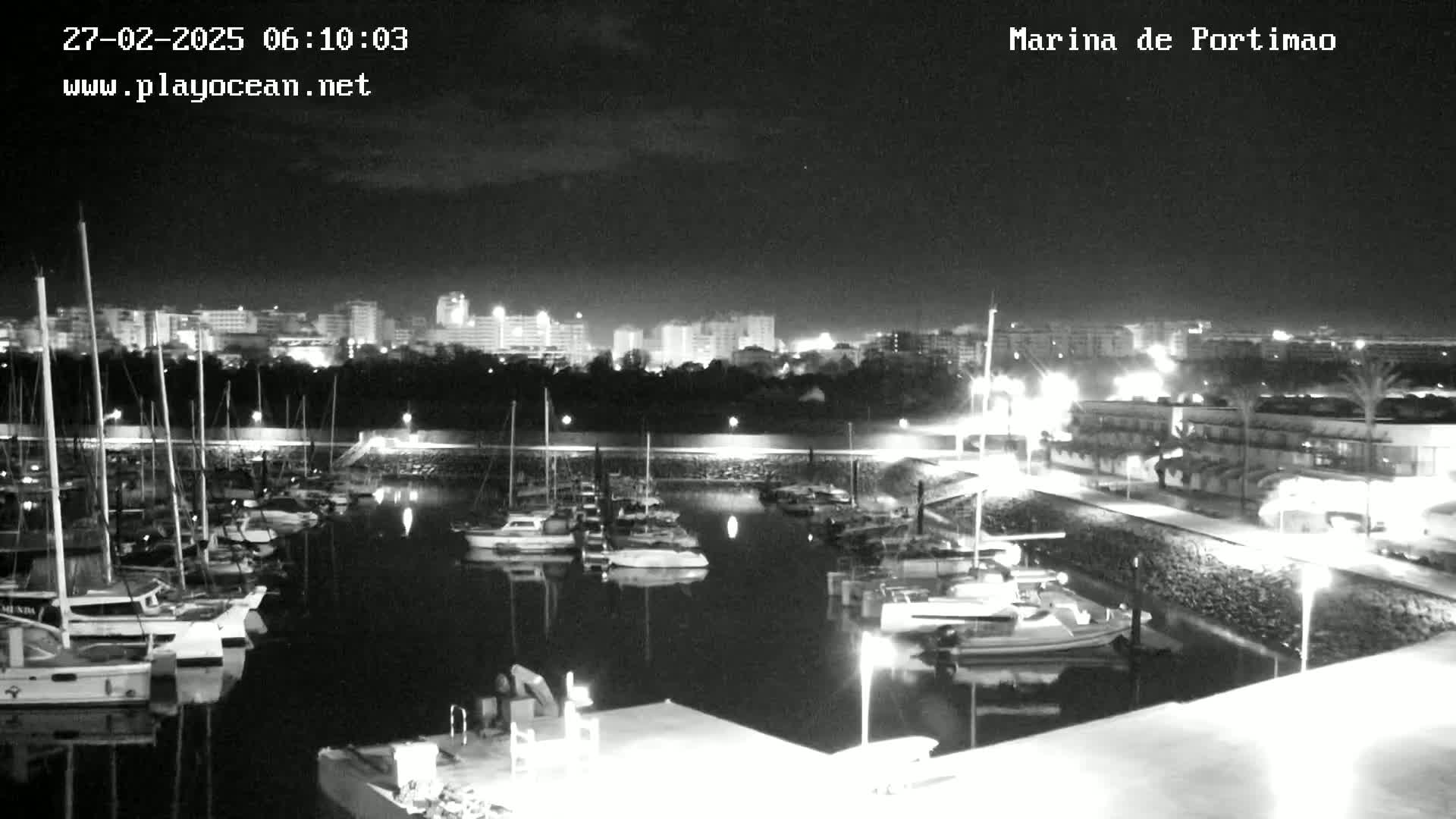 A nighttime, black and white view of a marina filled with sailboats, with a city skyline in the background under a partly cloudy sky.