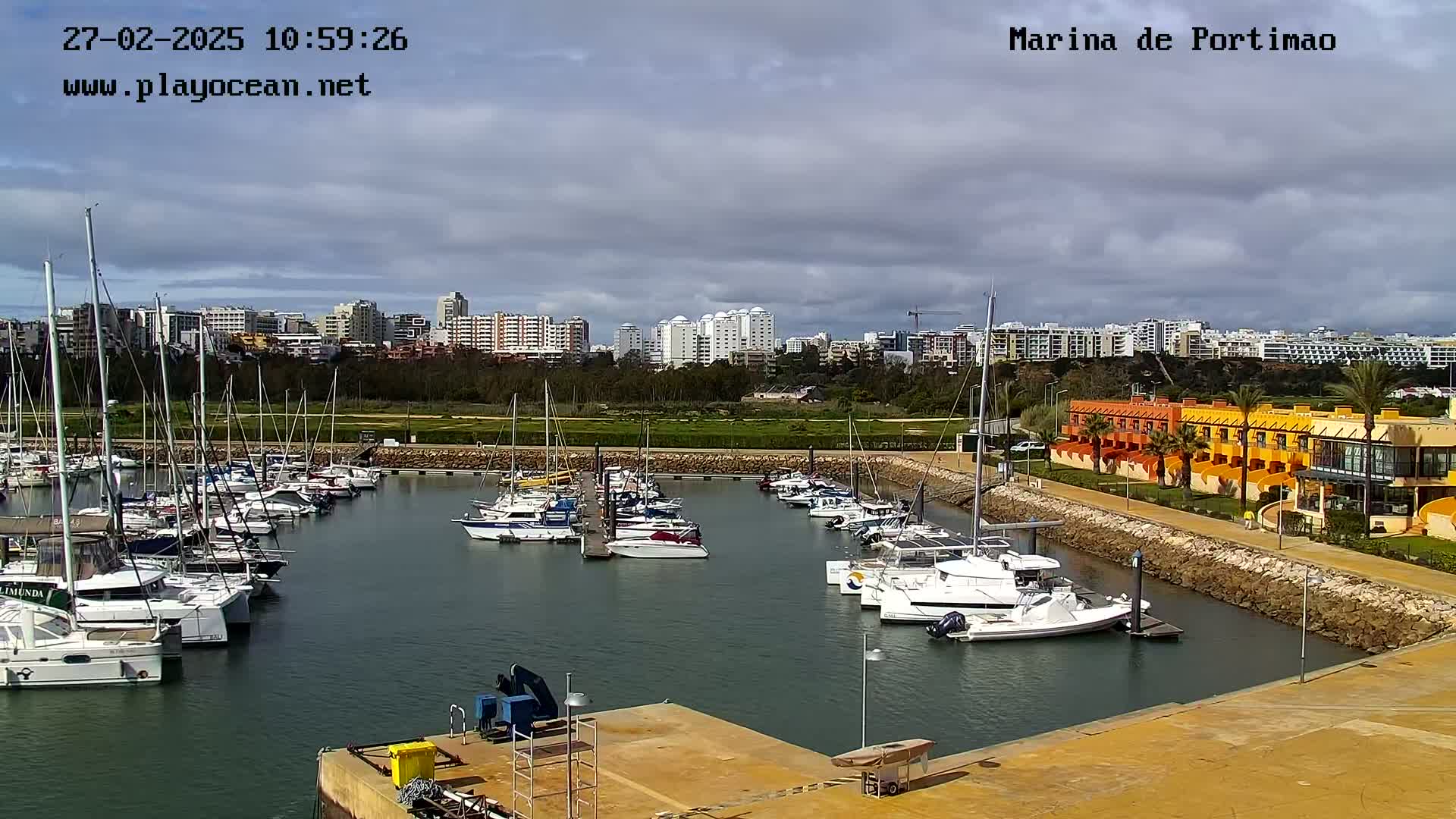 A marina filled with sailboats and motorboats is nestled beside a waterfront building complex, all under a partly cloudy sky.
