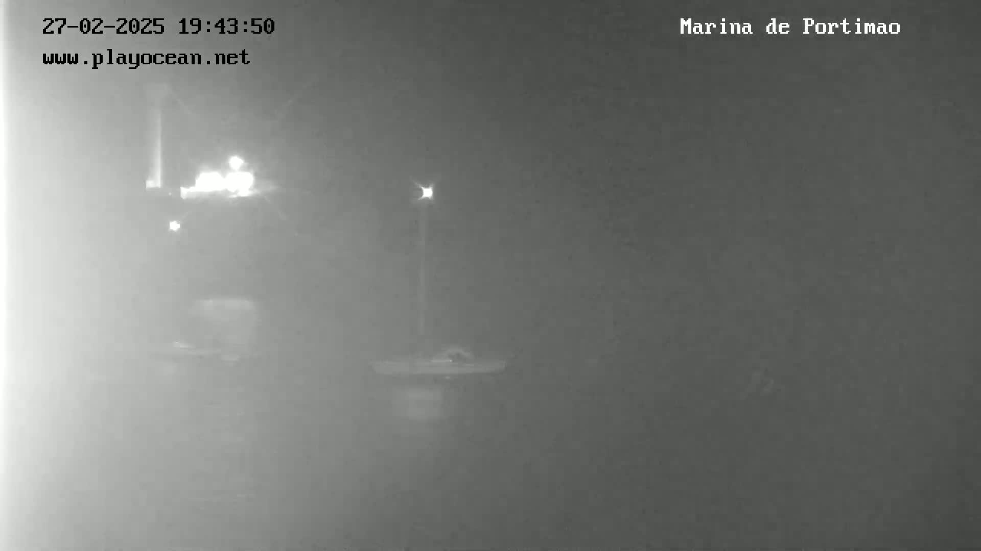 A nighttime, foggy view shows a dimly lit area with indistinct objects and faint lights.