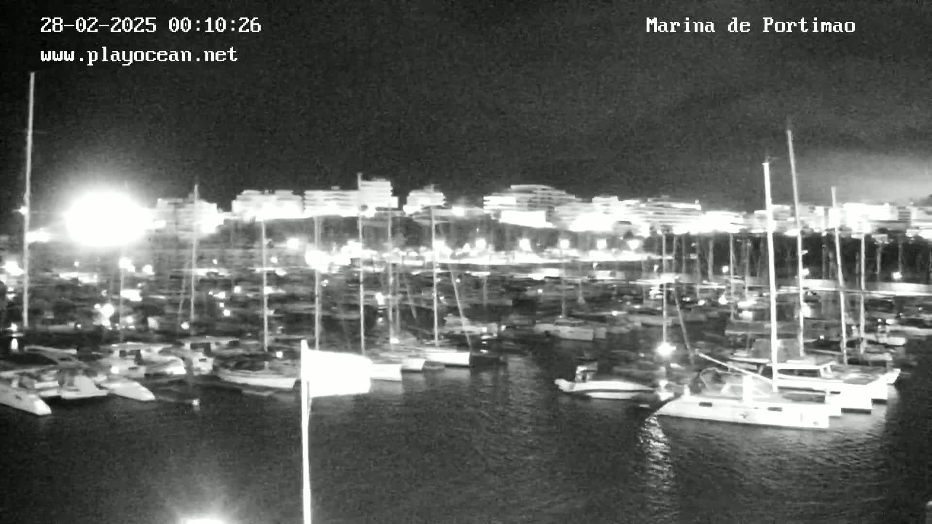 A nighttime view of a marina filled with numerous boats and yachts, illuminated by lights against a dark sky.