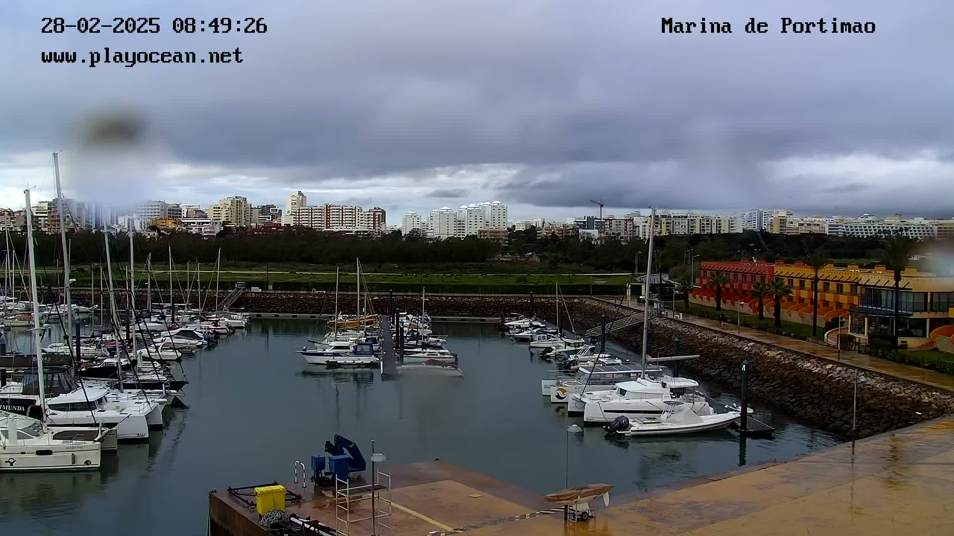 A marina filled with sailboats and motorboats is situated under a cloudy sky, with a shoreline of buildings and vegetation in the background.