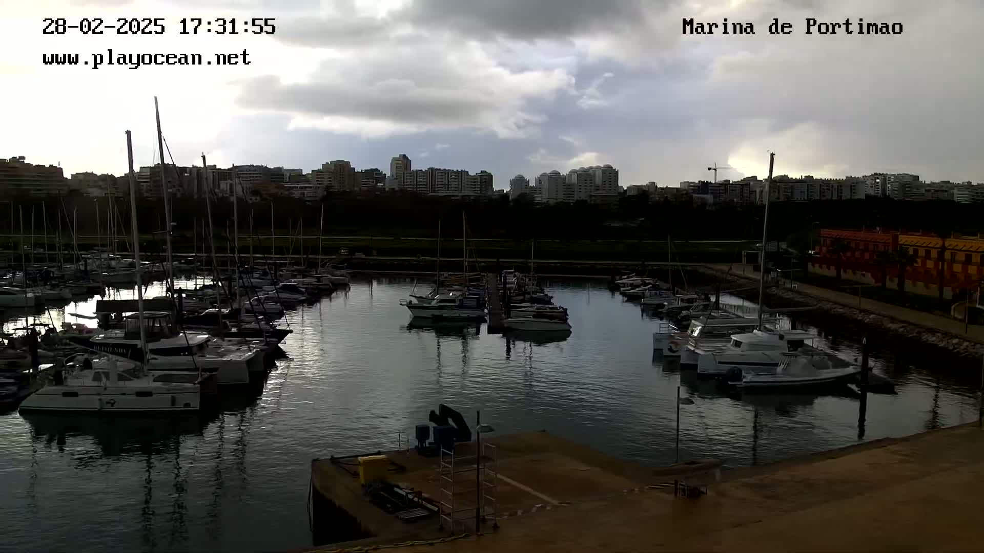 A marina filled with numerous sailboats and motorboats is situated under a cloudy sky near a city skyline.
