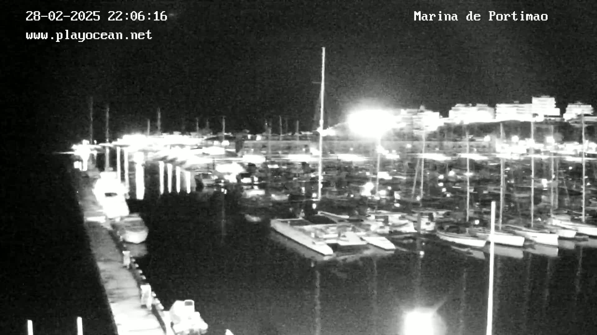 A nighttime, black-and-white view of a marina filled with numerous boats and yachts docked alongside a pier.