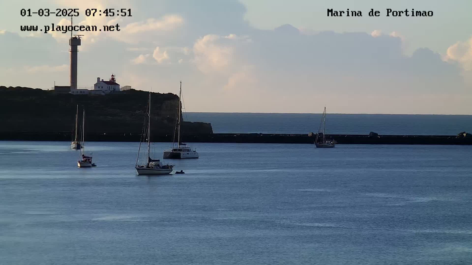 Several sailboats are moored in a calm harbor under a partly cloudy sky, with a lighthouse and tower visible on a nearby cliff.